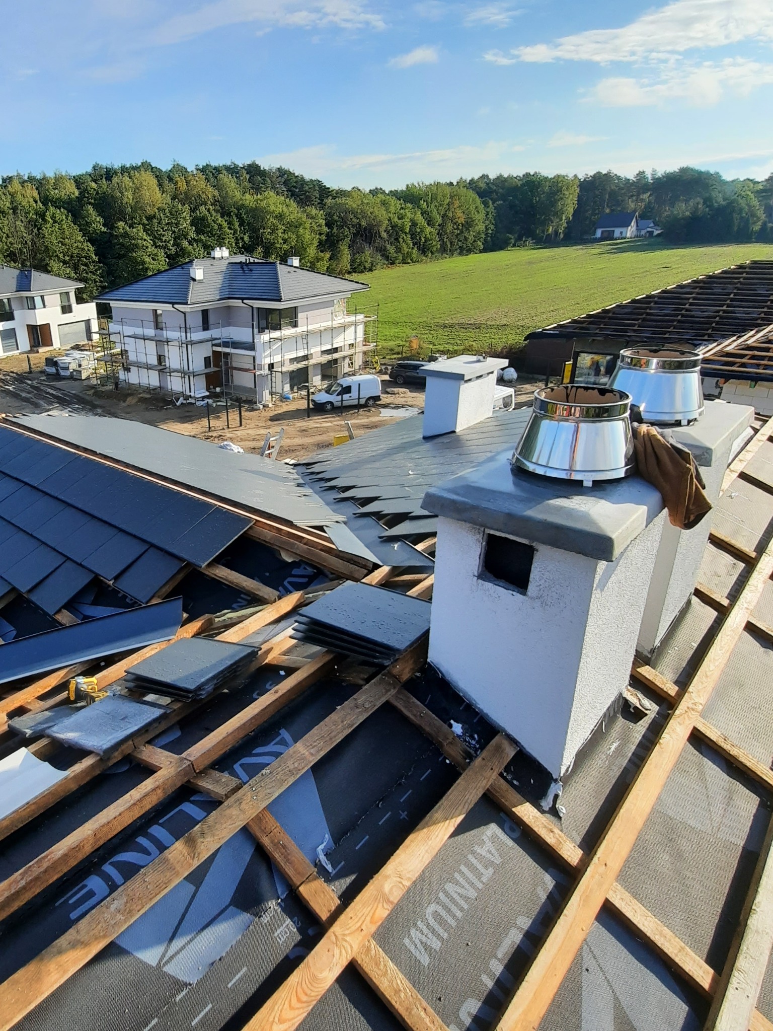 Montaż ceramicznej dachówki w trakcie budowy dachu, widoczne kominy z metalowymi nasadami, w tle dom w rusztowaniach i zielony krajobraz.