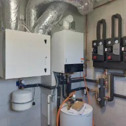 Instalacja rekuperacji z białymi obudowami Komfovent i rurami wentylacyjnymi pod sufitem, obok kotła Viessmann i pompy obiegowej Keller, na tle płytek ceramicznych.