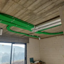 Zielone karbowane rury wentylacyjne przymocowane do surowego betonowego sufitu w budynku w stanie surowym, srebrne rury obok, widok przez okno na zalesione wzgórza.