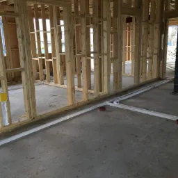 Wnętrze budynku w trakcie budowy z widoczną drewnianą konstrukcją szkieletową i ułożonymi na posadzce rurami kanalizacyjnymi z kształtkami.