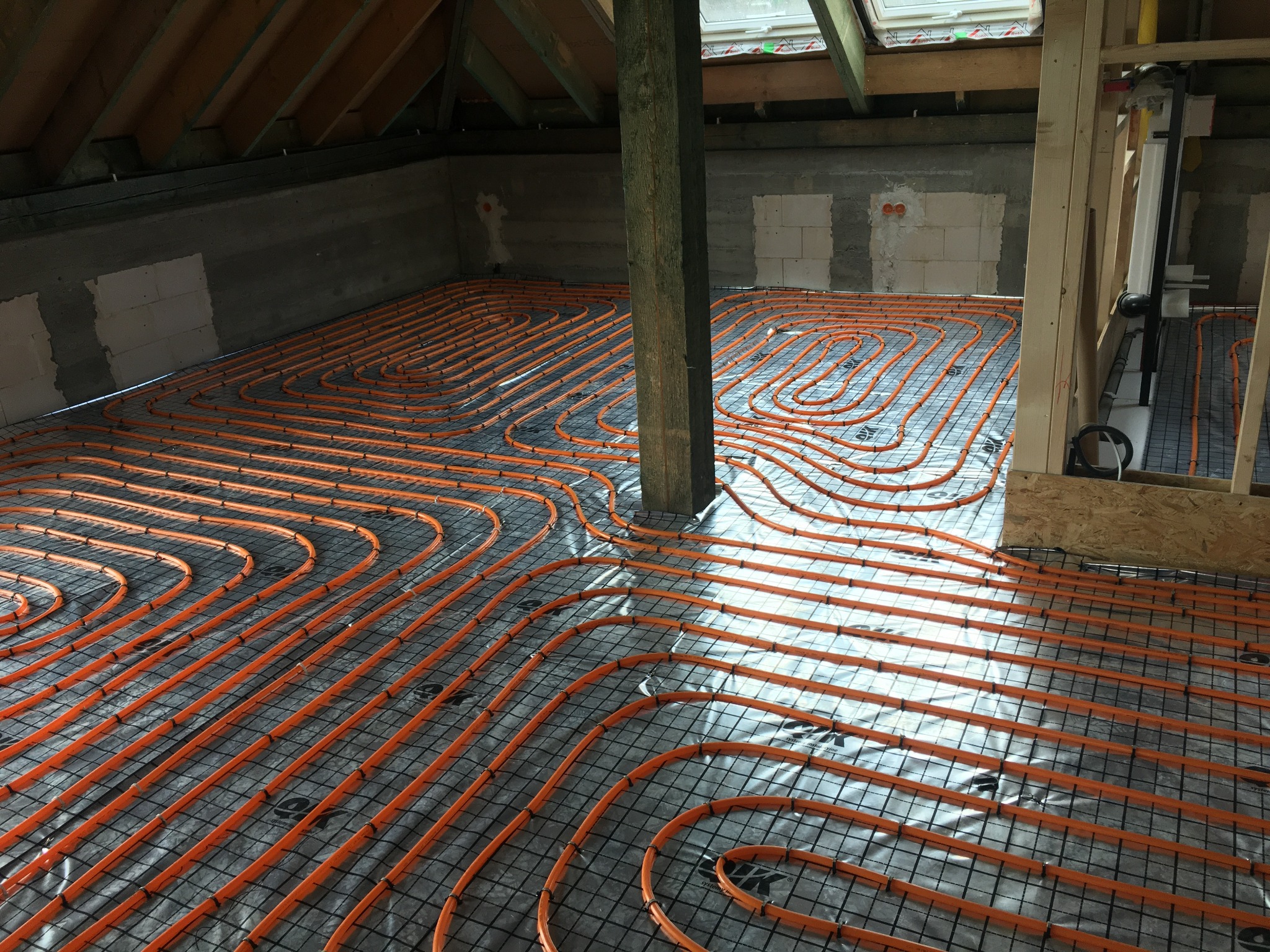 Pomarańczowe rury ogrzewania podłogowego ułożone na siatce w surowym wnętrzu poddasza z drewnianą konstrukcją dachu i pionowym słupem wspierającym, widoczne okno dachowe.