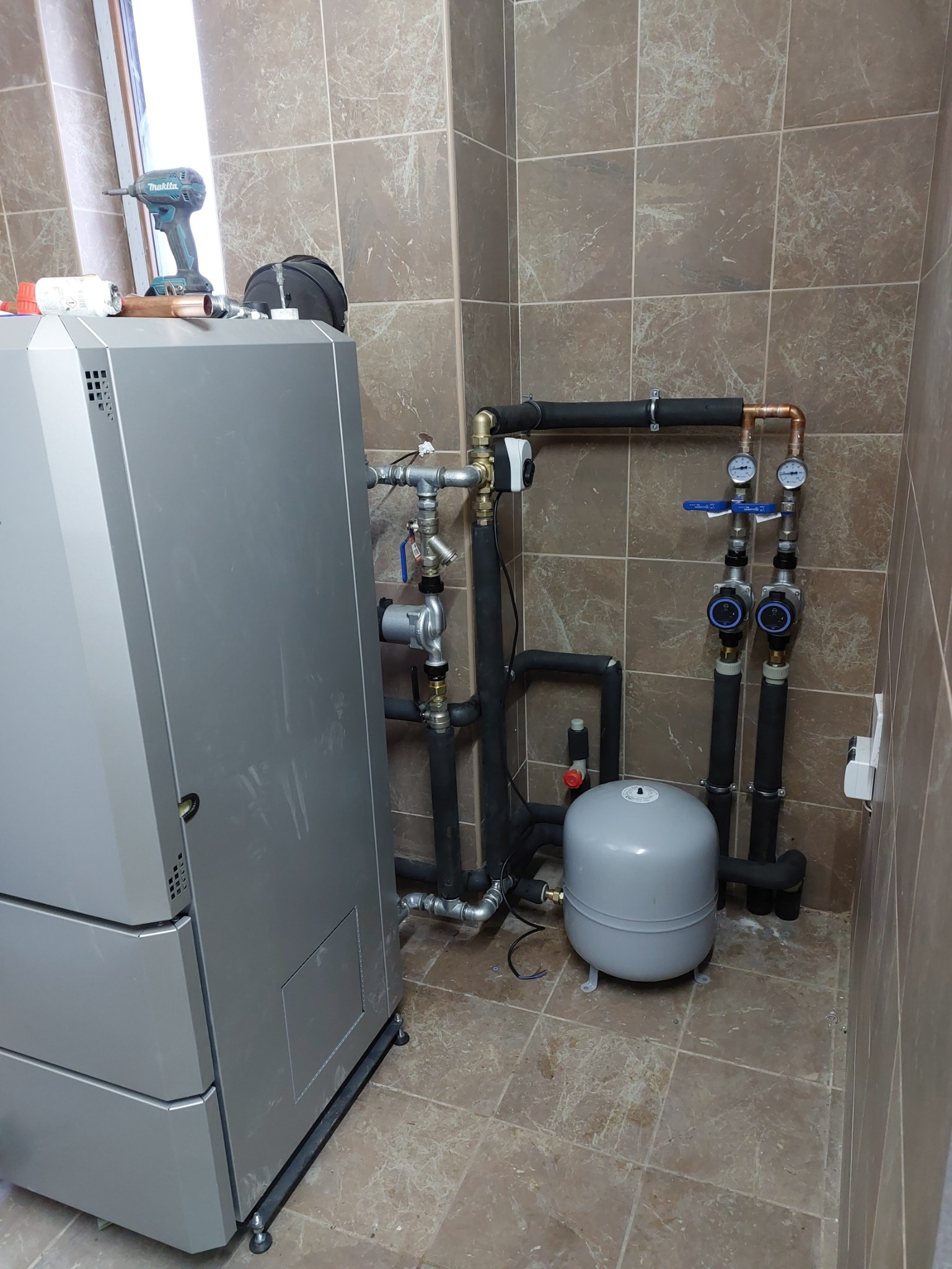 Szary piec grzewczy z podłączonymi rurami, manometrami i pompami obiegowymi w pomieszczeniu wyłożonym brązowymi płytkami, w tle wiertarka akumulatorowa na piecu.