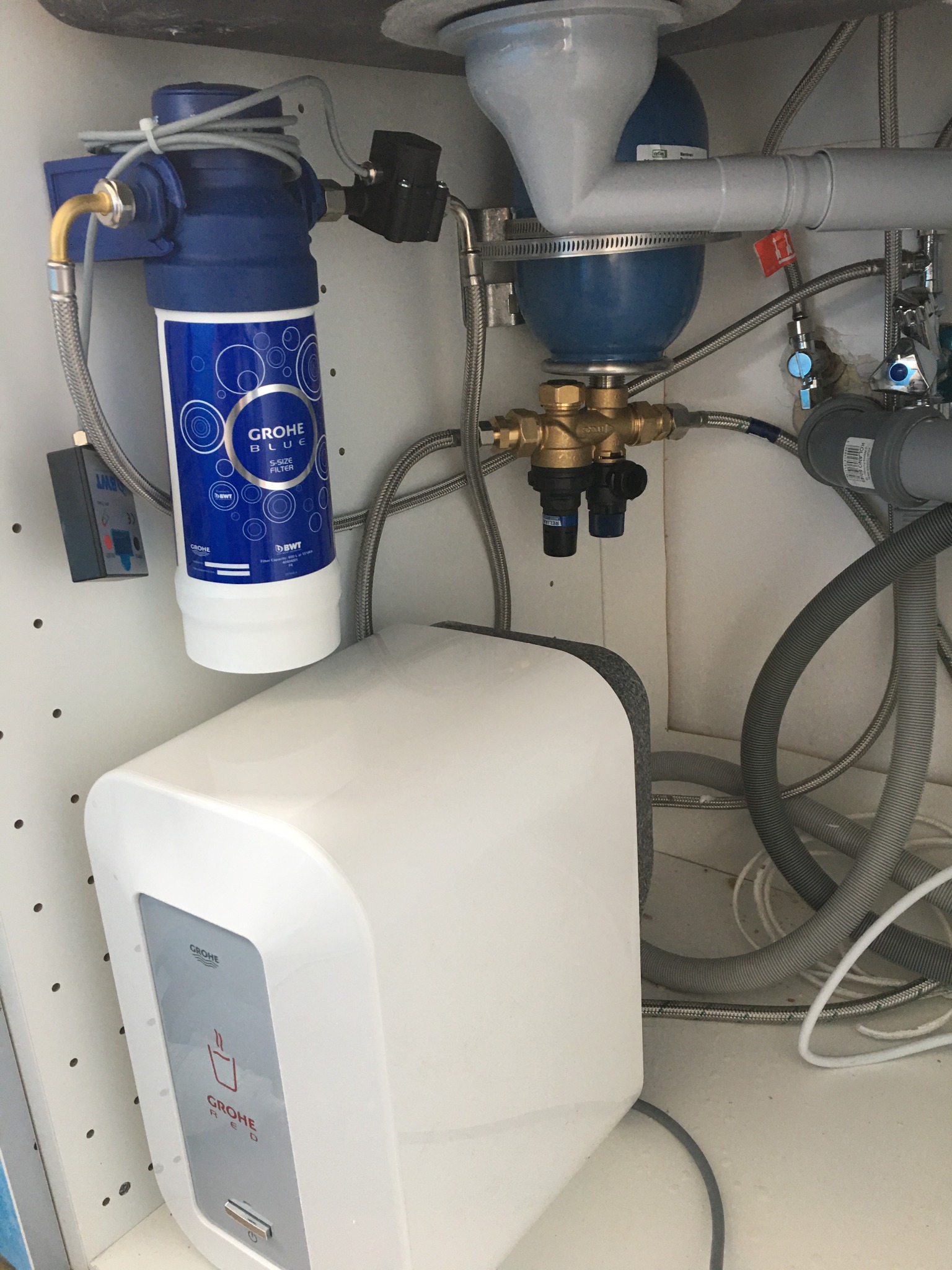 Instalacja pod zlewem z filtrem wody Grohe Blue, zbiornikiem ciśnieniowym, zaworami i rurami doprowadzającymi wodę, widoczne elementy instalacji elektrycznej.