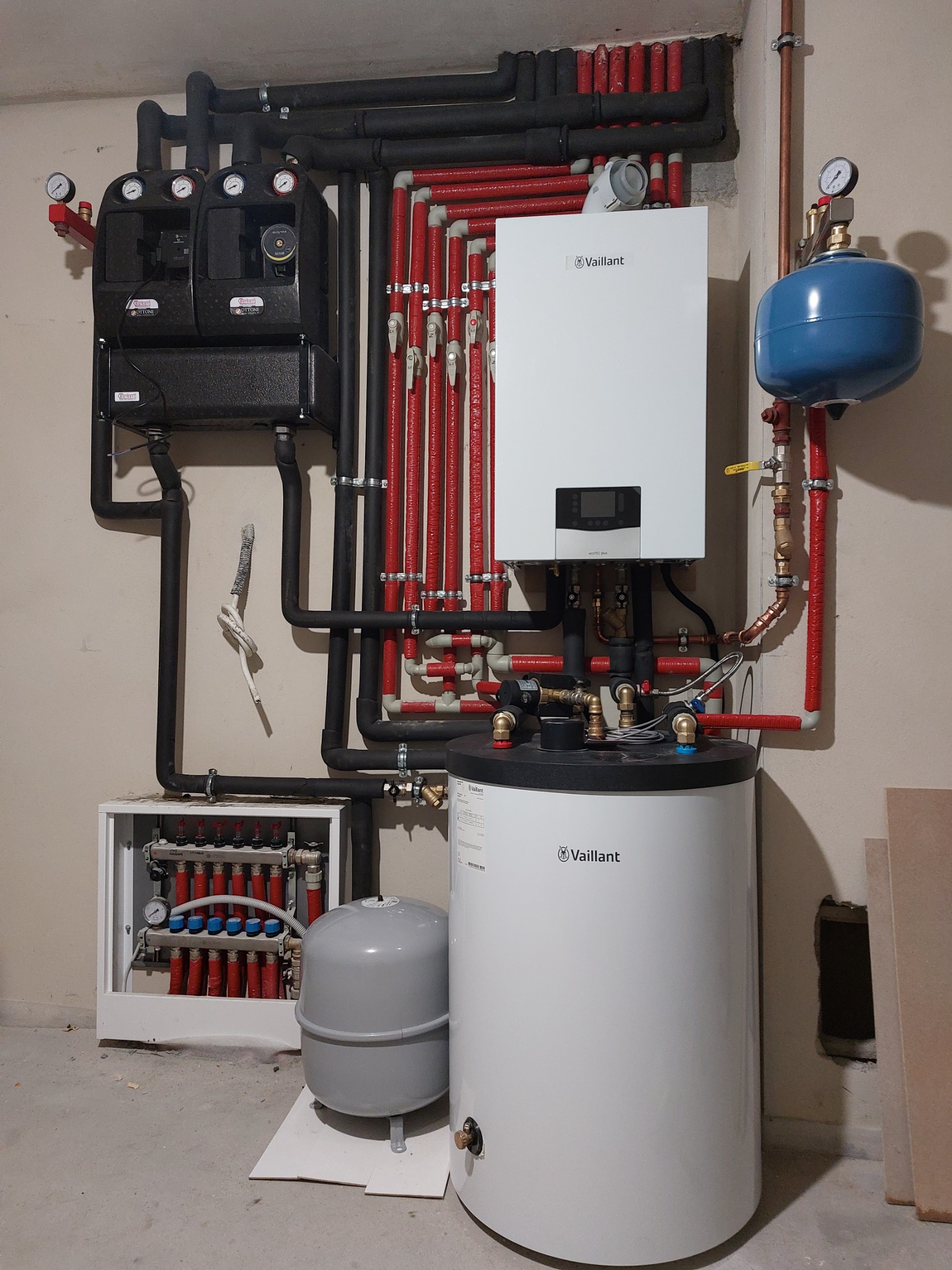Kompletna instalacja grzewcza w kotłowni: biały piec Vaillant, zasobnik ciepłej wody, rozdzielacz z licznymi rurami w izolacji termicznej, czarne pompy obiegowe. Widoczne manometry i zawory.