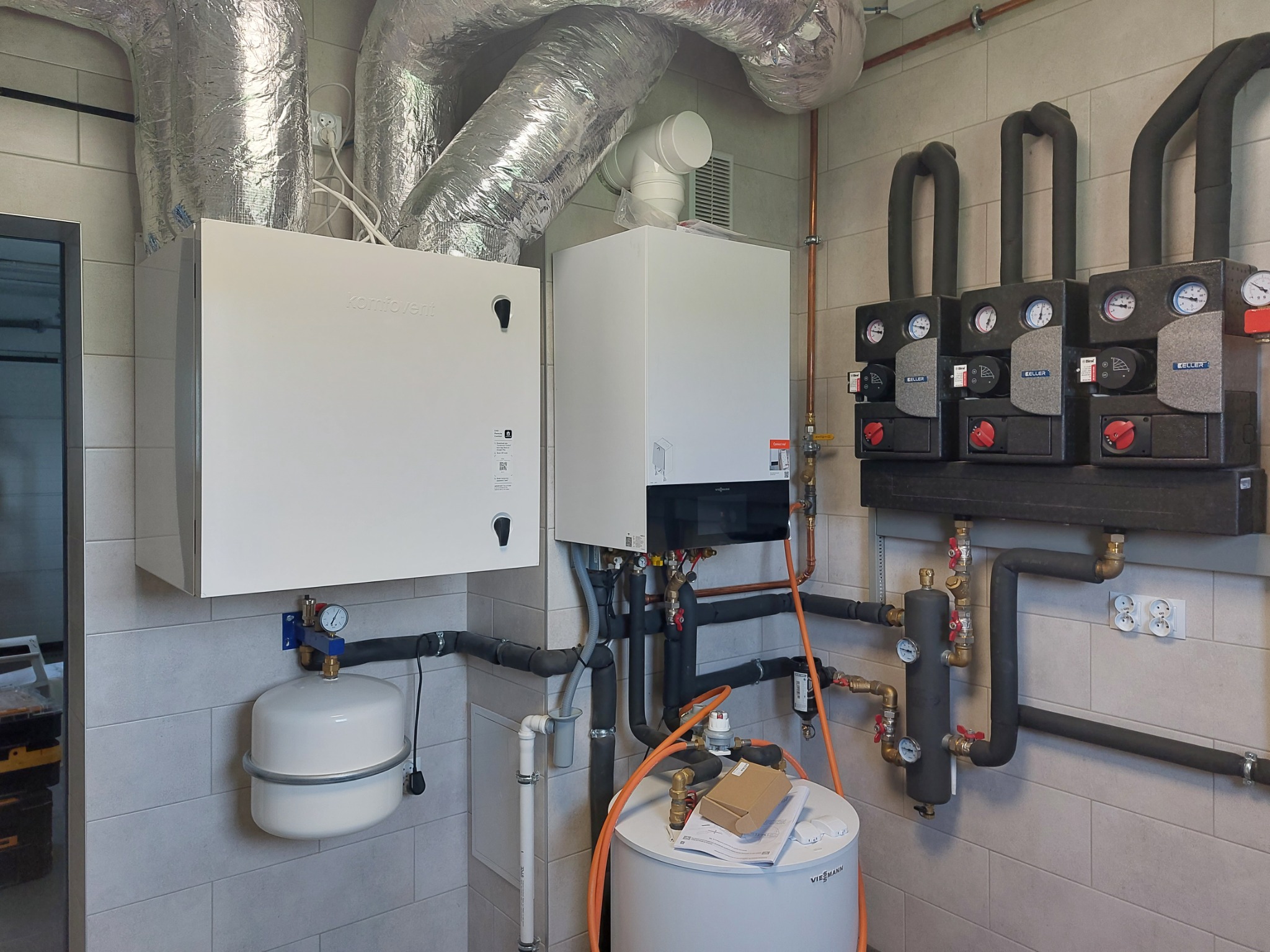 Instalacja rekuperacji z białymi obudowami Komfovent i rurami wentylacyjnymi pod sufitem, obok kotła Viessmann i pompy obiegowej Keller, na tle płytek ceramicznych.