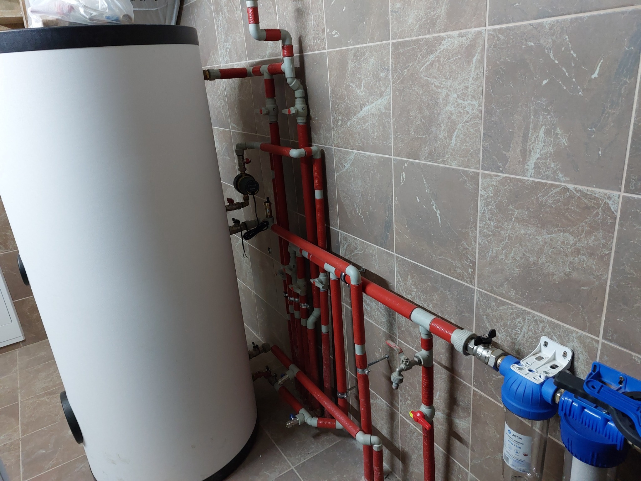 Biały zasobnik ciepłej wody użytkowej z podłączonymi czerwonymi rurami instalacji grzewczej i filtrami wody na tle brązowych płytek ściennych.