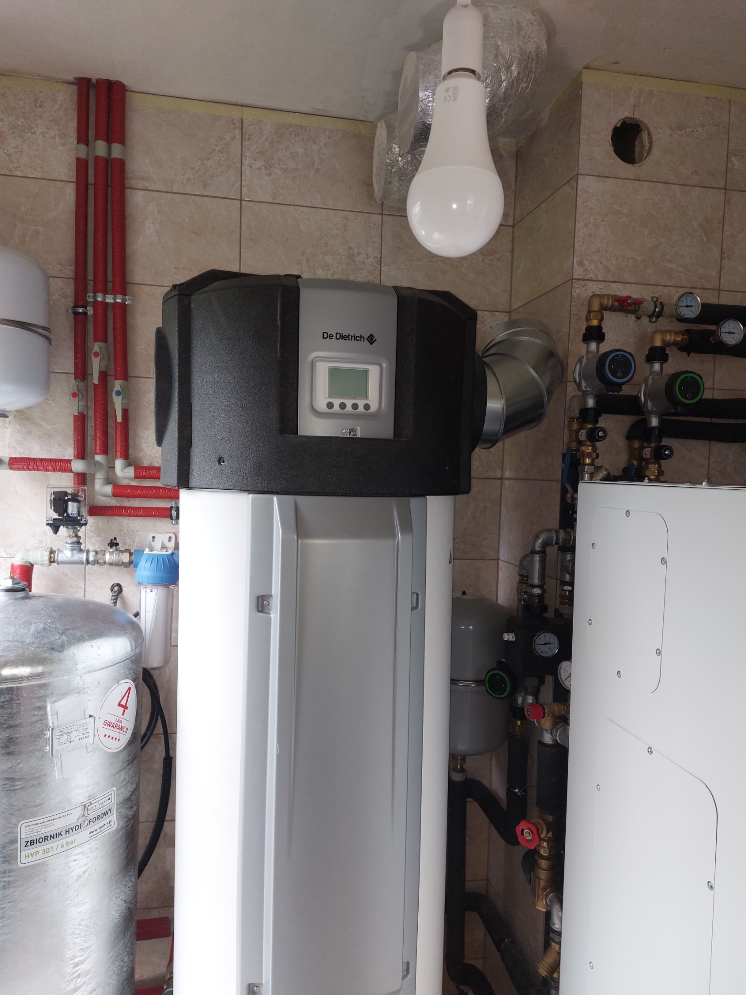 Zamontowany hydrofor De Dietrich z widocznymi rurami, zaworami i manometrami w pomieszczeniu z beżowymi płytkami, oświetlony wiszącą żarówką LED.