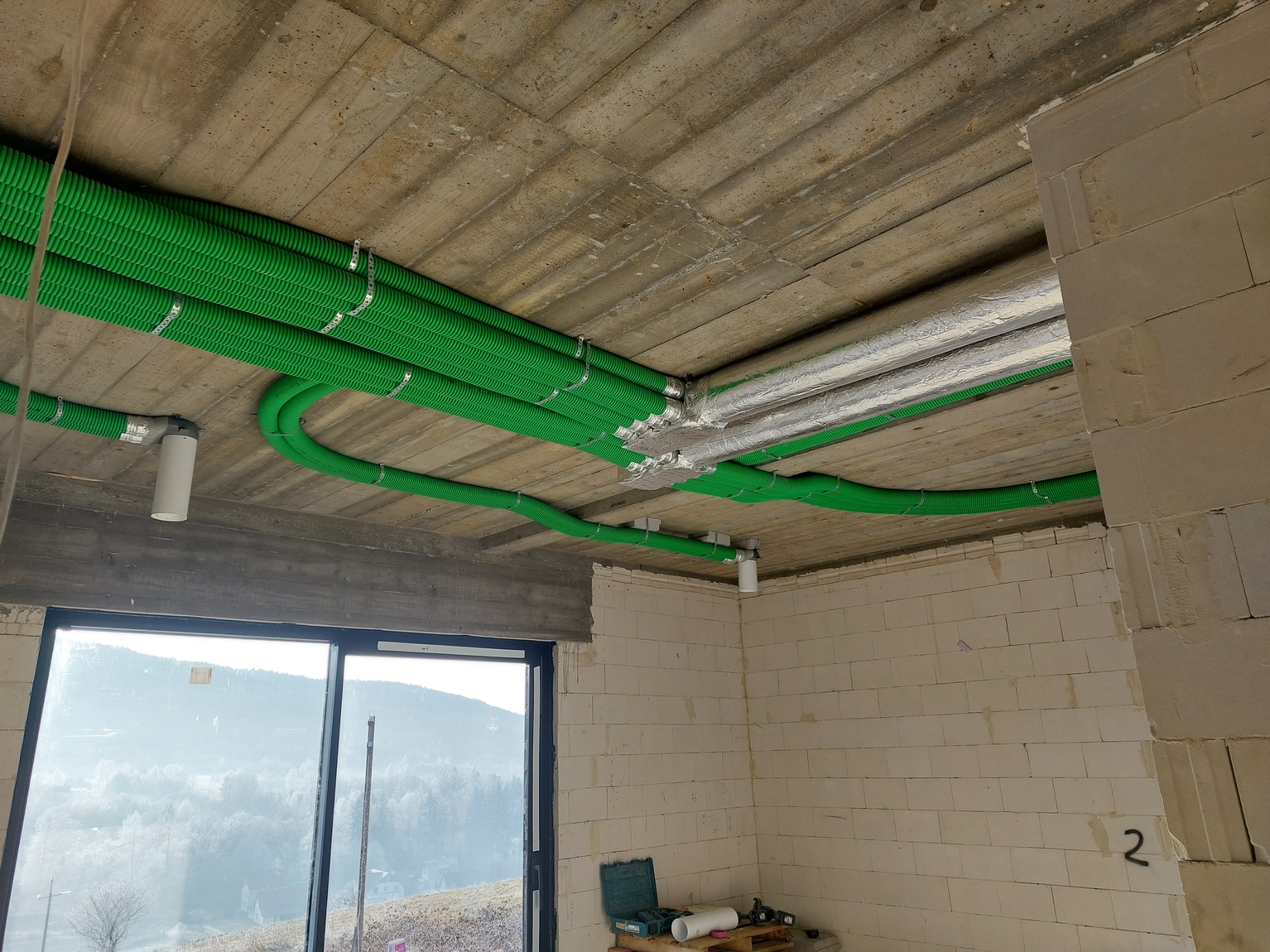 Zielone karbowane rury wentylacyjne przymocowane do surowego betonowego sufitu w budynku w stanie surowym, srebrne rury obok, widok przez okno na zalesione wzgórza.