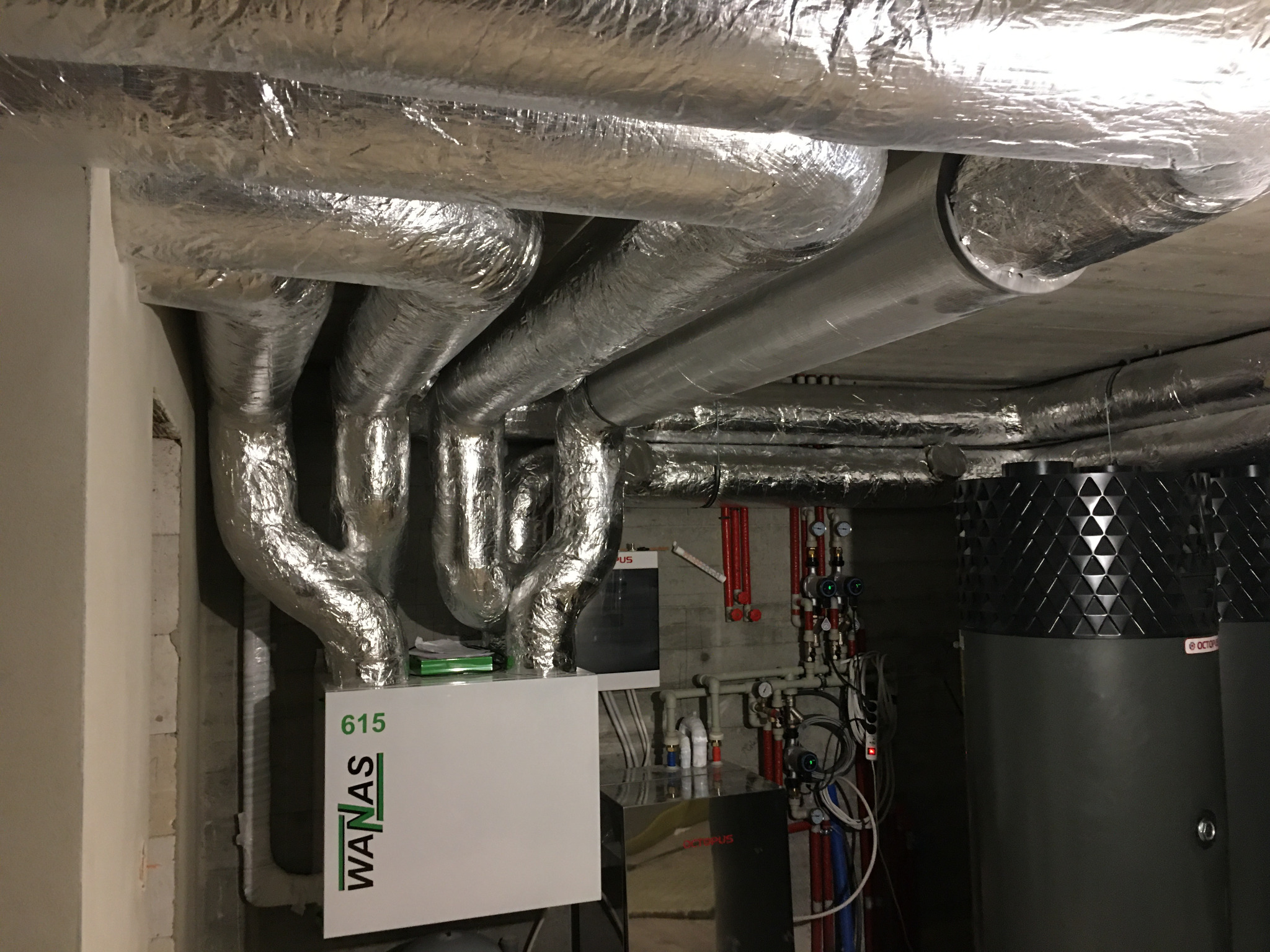 System kanałów wentylacyjnych z folii aluminiowej pod sufitem, połączony z białą jednostką WANAS 615, widoczny również system rur i zbiorniki Octopus, w pomieszczeniu technicznym.