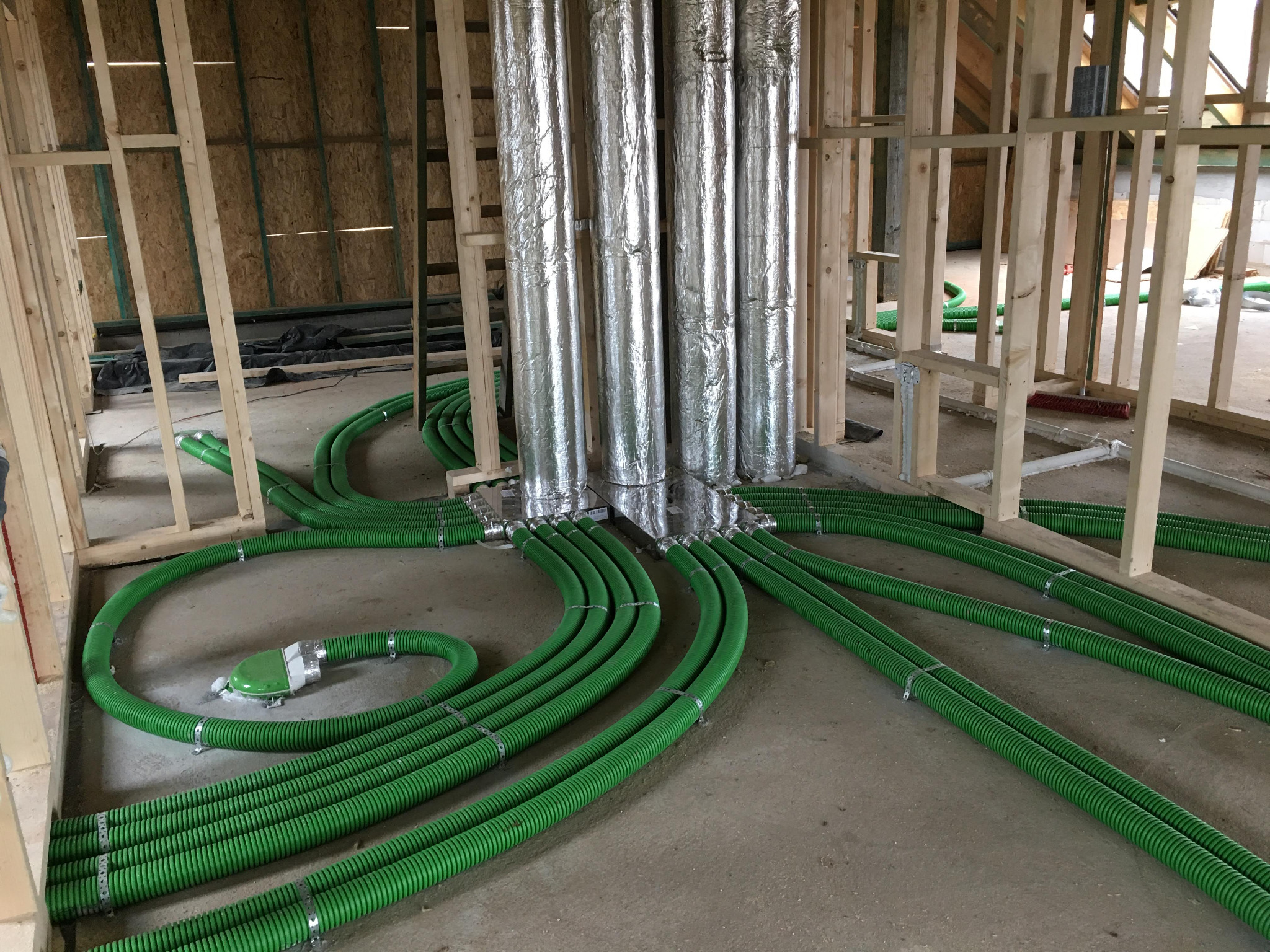 Wnętrze w trakcie budowy w Żywcu z widocznymi elementami instalacji wentylacyjnej: srebrne, zaizolowane pionowe rury oraz zielone, karbowane przewody wentylacyjne rozchodzące się po podłodze.