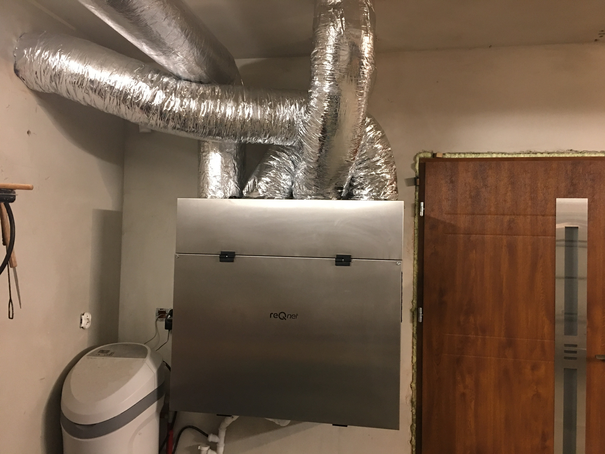 Srebrny rekuperator z widocznymi przewodami wentylacyjnymi w izolacji, zamontowany w pomieszczeniu technicznym obok zmiękczacza wody i drzwi z ościeżnicą uszczelnioną pianką montażową.