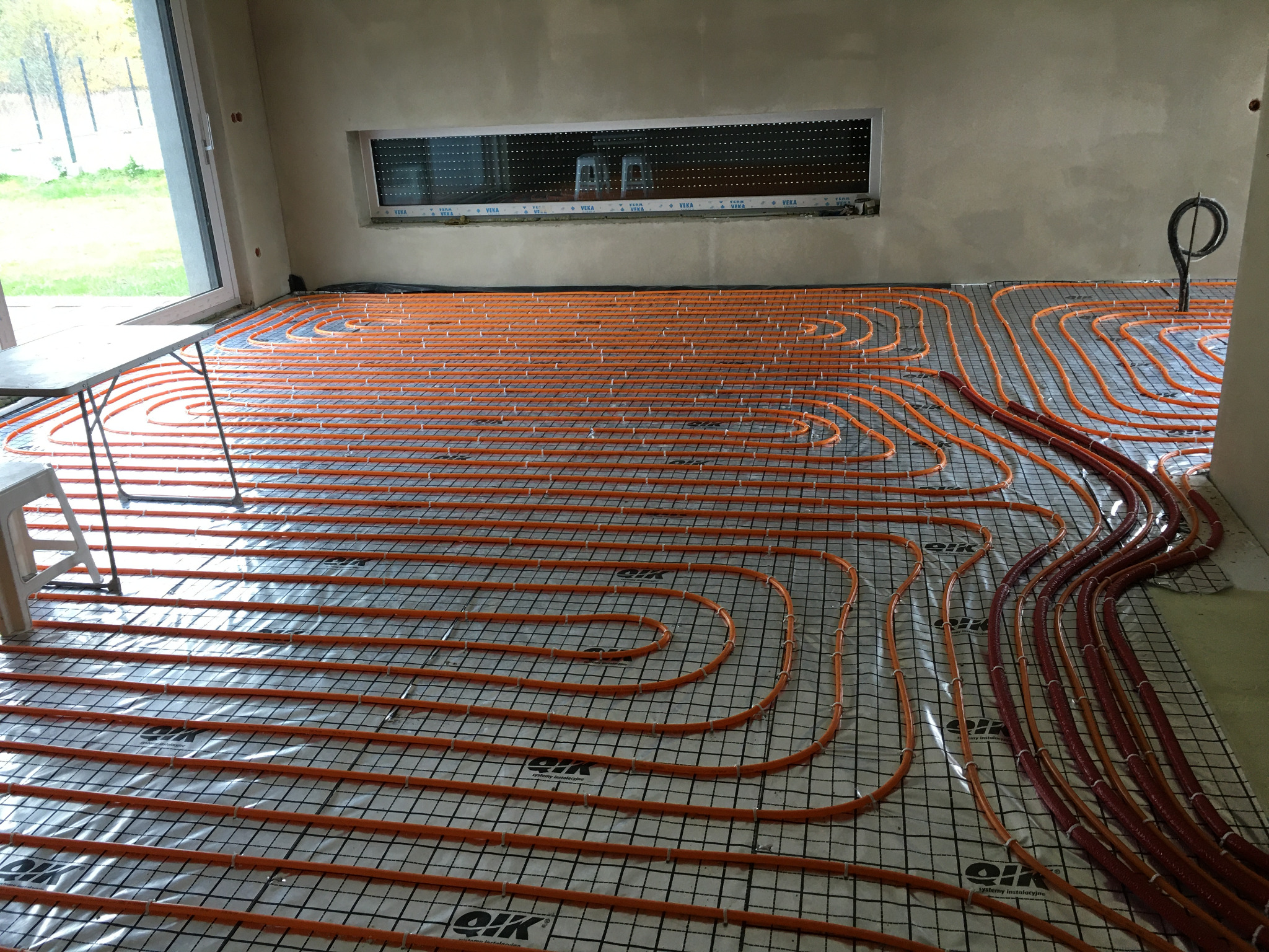 Pomarańczowe rury ogrzewania podłogowego ułożone na izolacji z siatką w surowym wnętrzu, widoczne okno z roletami i fragment krajobrazu za nim.
