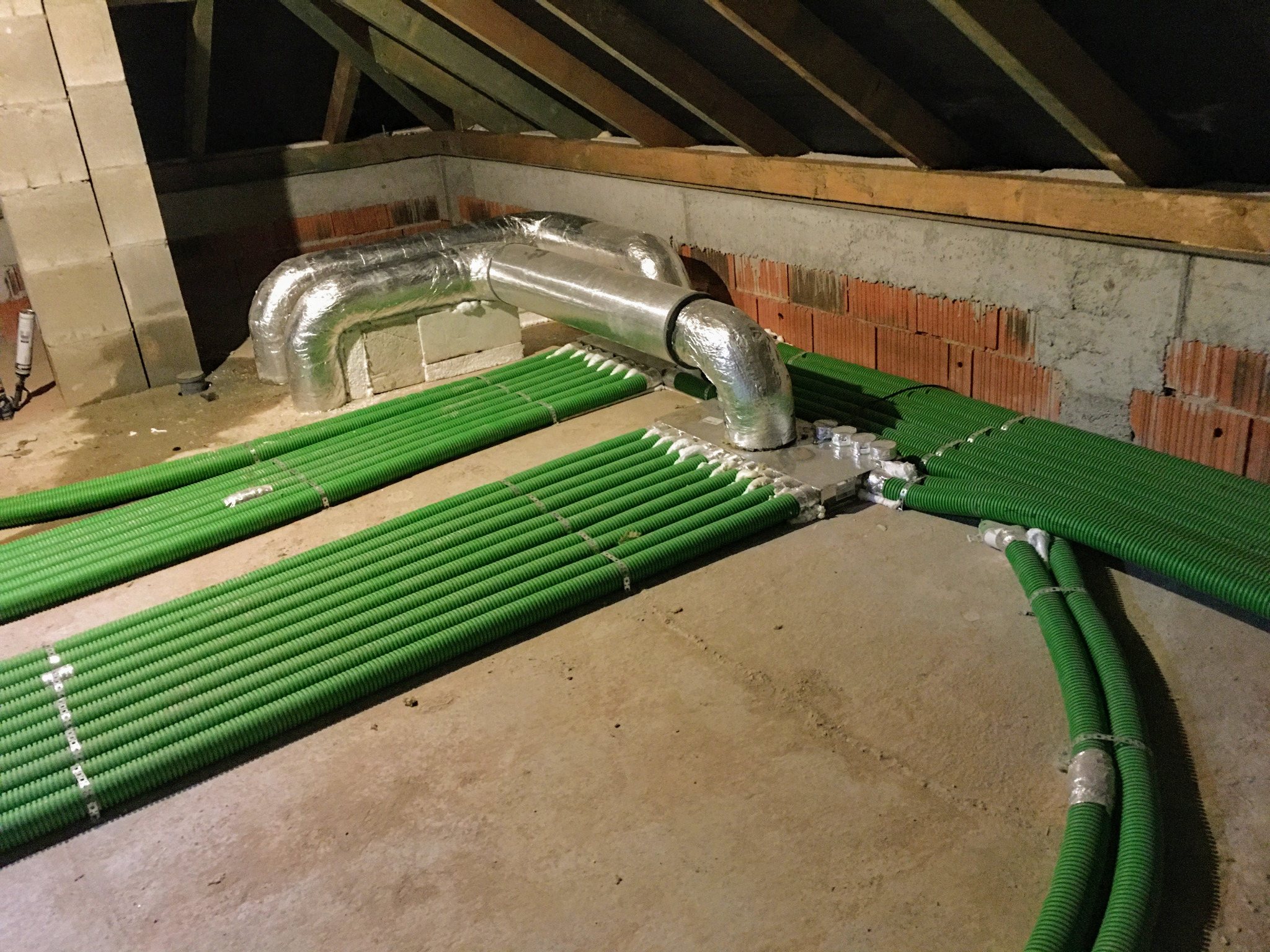 Rozprowadzenie systemu wentylacyjnego z zielonych rur karbowanych w przestrzeni poddasza, widoczne izolowane aluminiową folią rury i elementy łączeniowe na tle muru z cegieł i drewnianej...