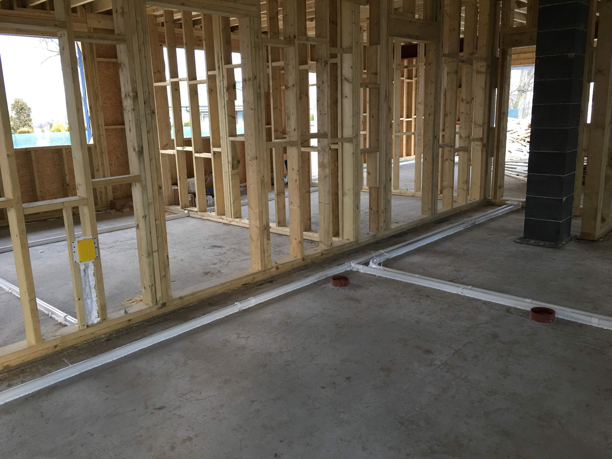 Wnętrze budynku w trakcie budowy z widoczną drewnianą konstrukcją szkieletową i ułożonymi na posadzce rurami kanalizacyjnymi z kształtkami.