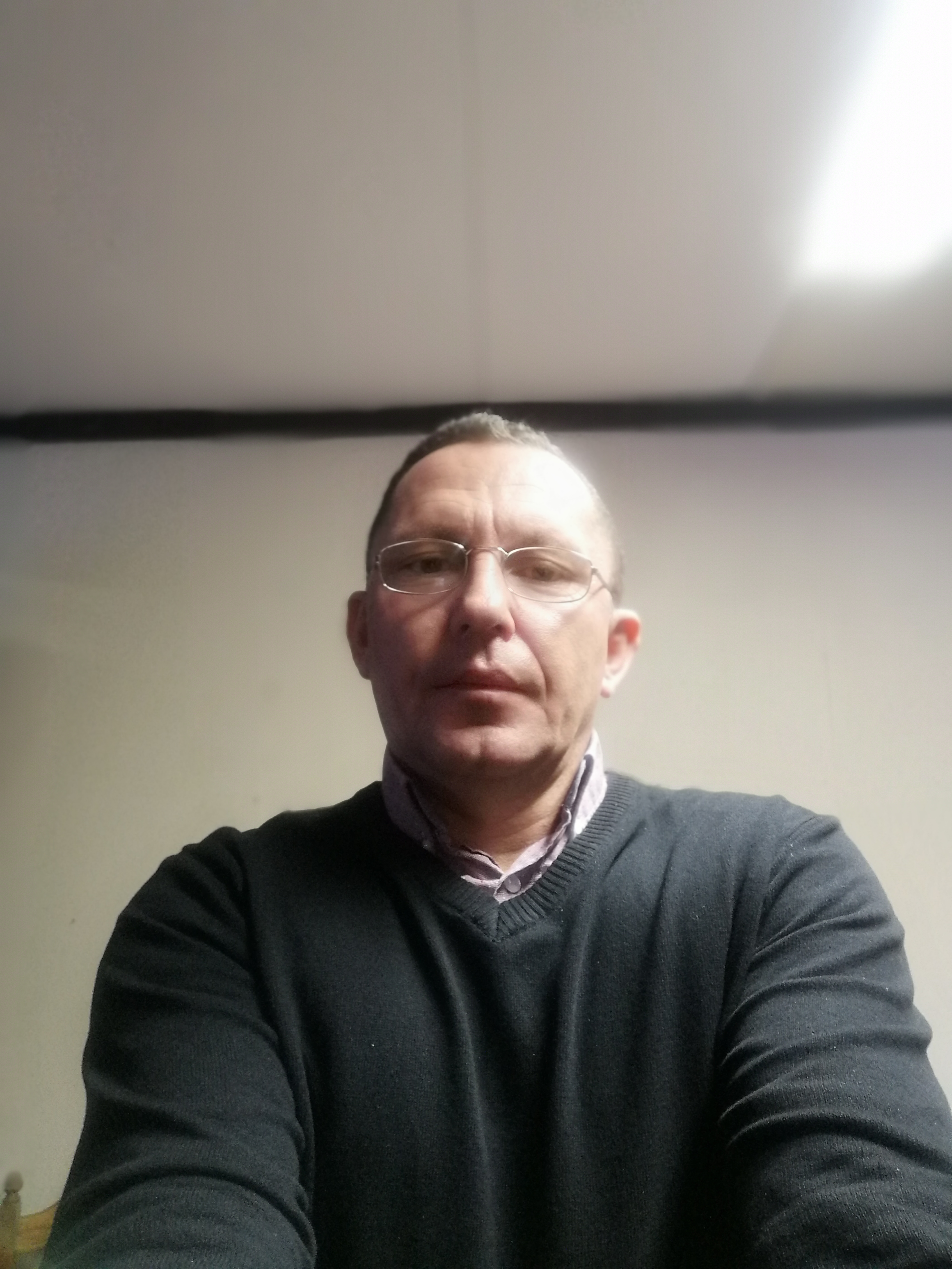 Portret mężczyzny w okularach i ciemnym swetrze na tle jasnej ściany z widocznym fragmentem sufitu.