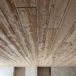 Drewniany sufit wyłożony jasnymi, lakierowanymi deskami z wyraźnym usłojeniem, widziany pod kątem, z fragmentem surowej ściany w tle.