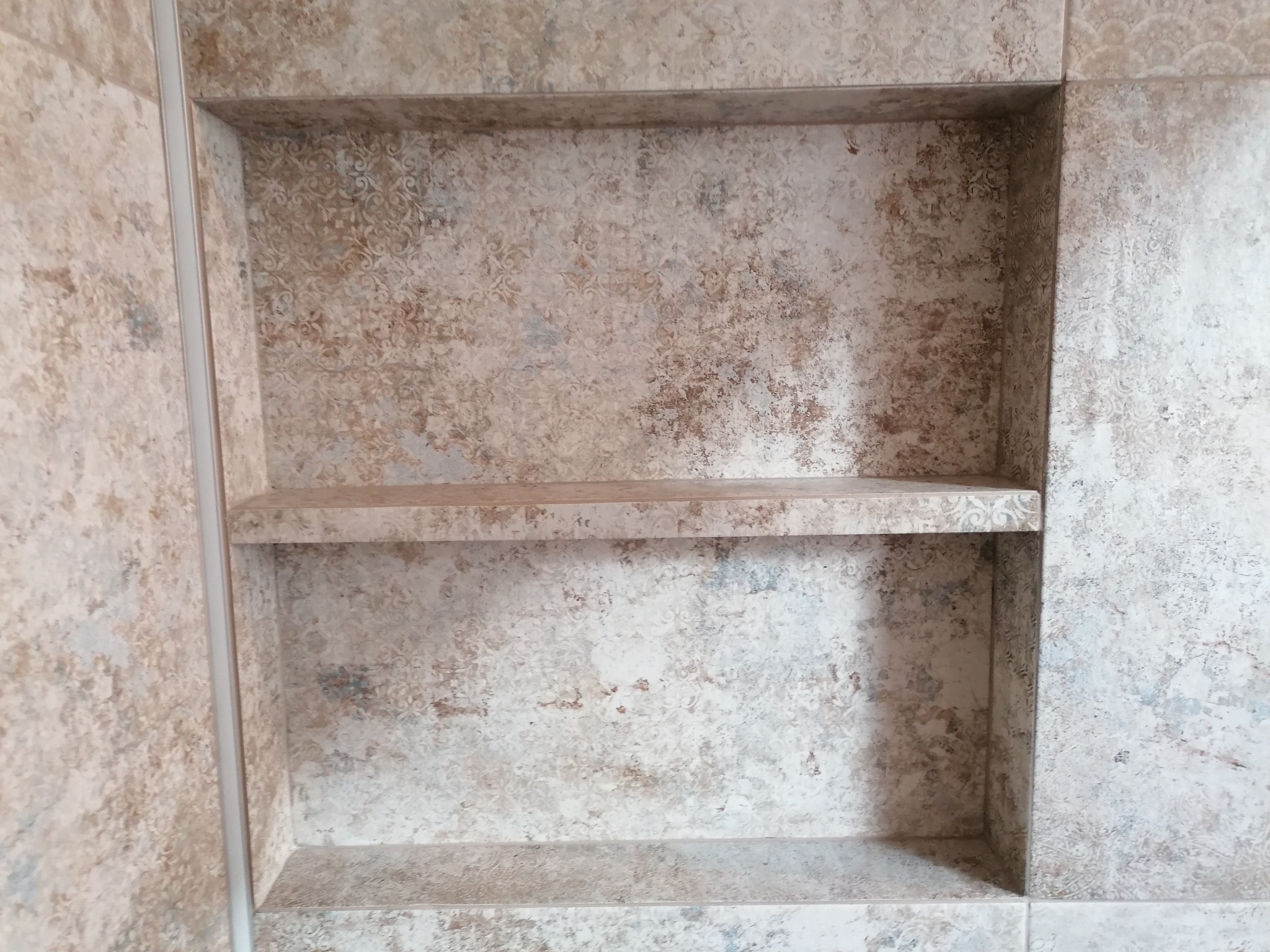 Wnęka z półką wyłożona płytkami o wzorze imitującym starą mozaikę w odcieniach beżu i szarości, widoczna fuga między płytkami.