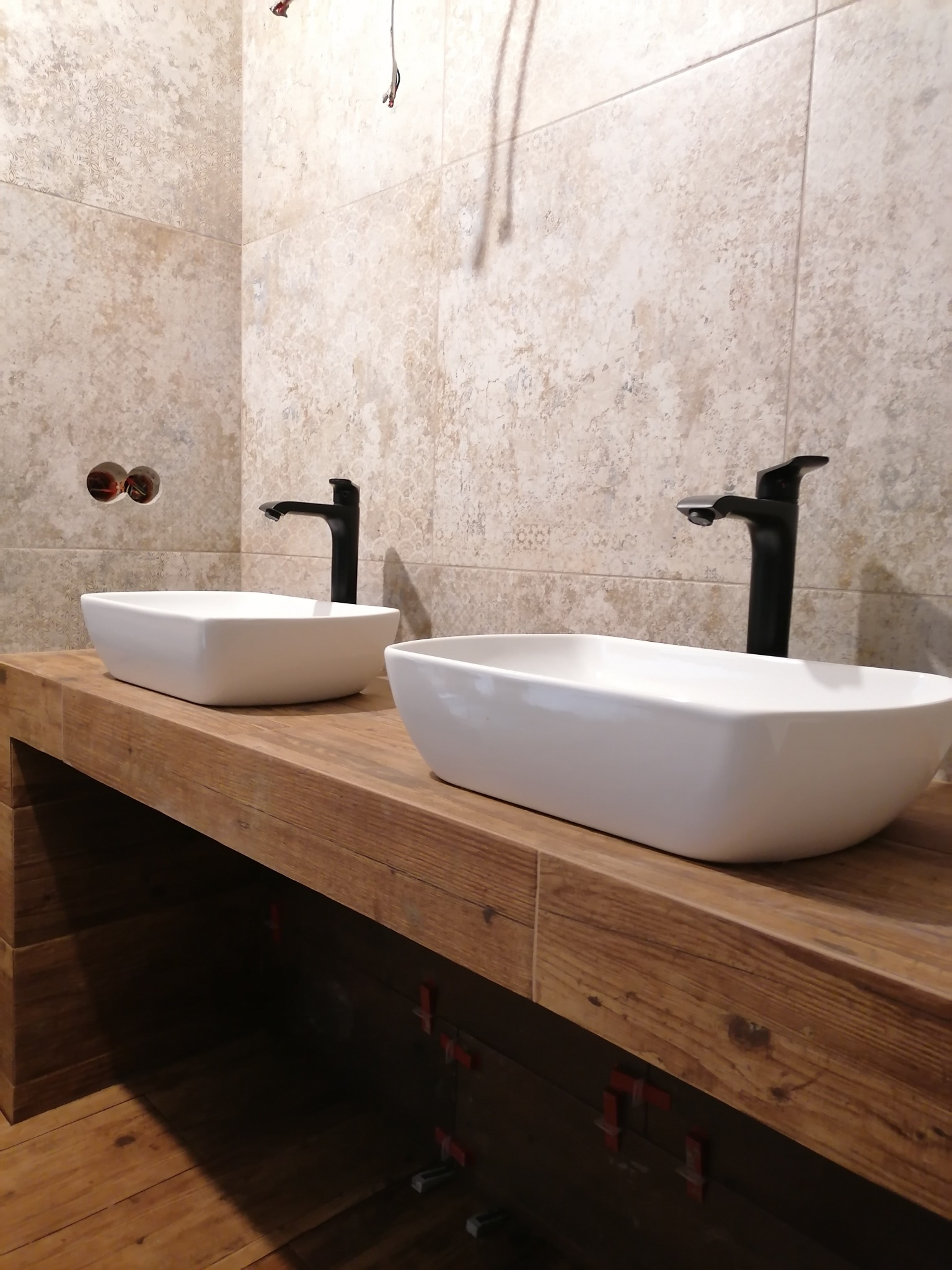 Dwie białe, prostokątne umywalki nablatowe z czarnymi bateriami na blacie z drewna o ciepłym odcieniu, tło stanowią beżowe płytki ścienne z delikatnym wzorem, widoczne elementy instalacji...