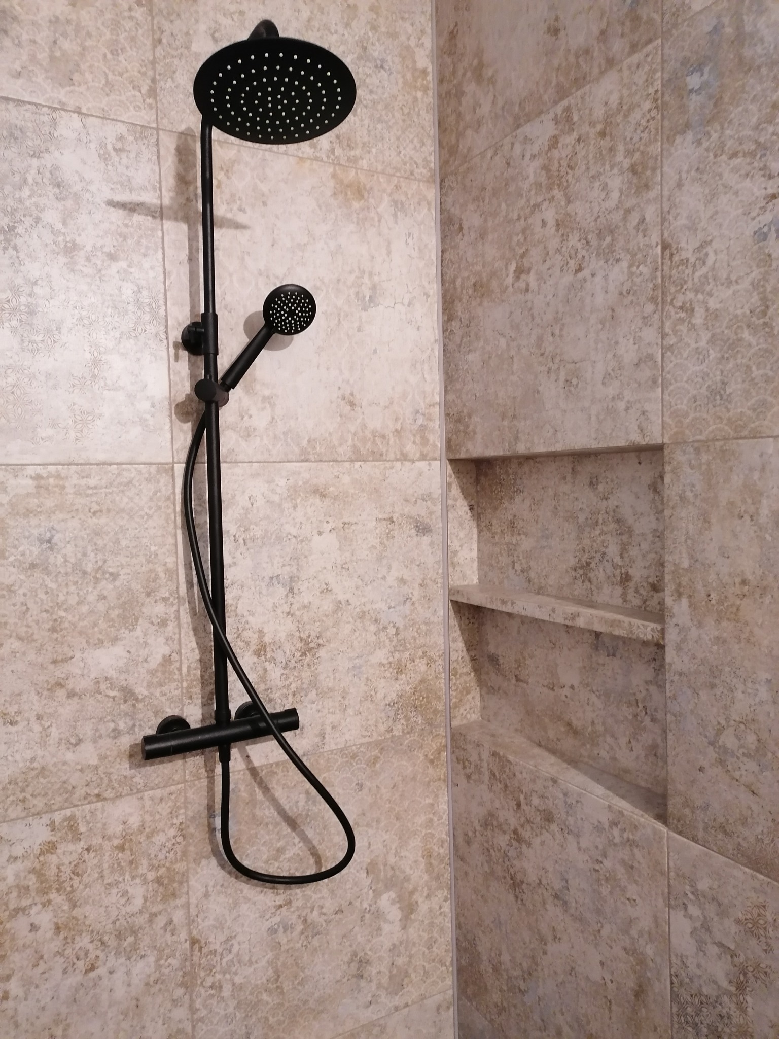 Zamontowany czarny zestaw prysznicowy z deszczownicą i słuchawką na tle beżowych płytek imitujących kamień z wnęką z półkami.