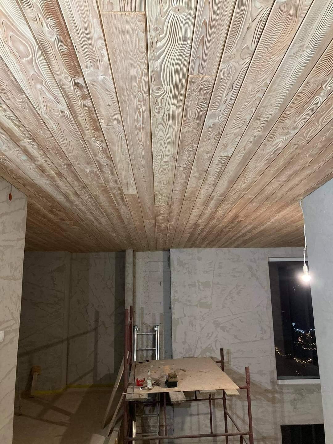 Drewniany sufit z jasnych desek z wyraźnym usłojeniem, widok z dołu, w tle surowe ściany i rusztowanie.