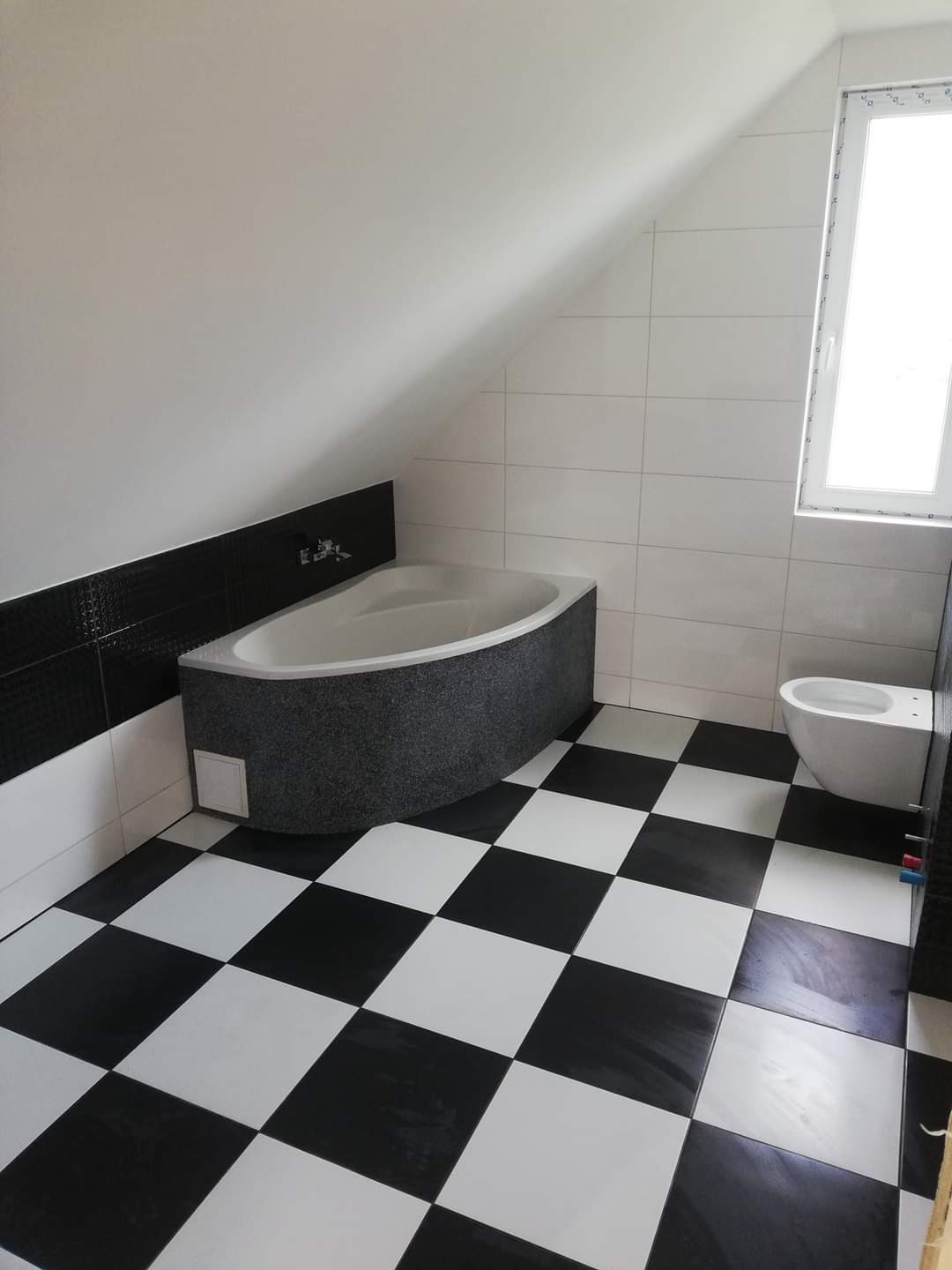 Czarno-biała łazienka na poddaszu z wanną obudowaną szarą mozaiką, białą toaletą i podłogą w szachownicę.