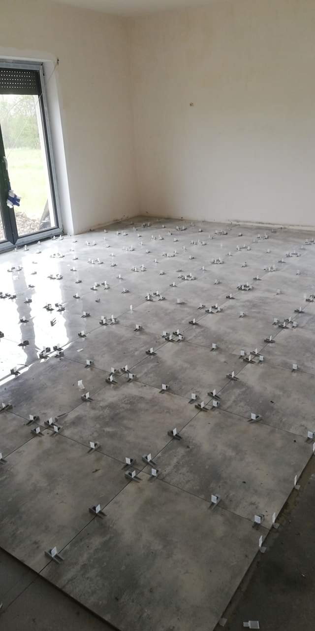 Szare, błyszczące płytki podłogowe w trakcie układania, z widocznymi krzyżykami dystansowymi i sznurkami do wyznaczania linii, w jasnym pomieszczeniu z oknem.