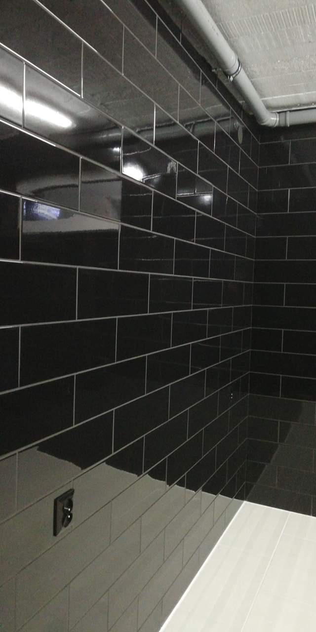 Ściana wyłożona błyszczącymi, czarnymi płytkami w kształcie cegiełek z szarą fugą, widoczny fragment białej podłogi i szara rura pod sufitem.