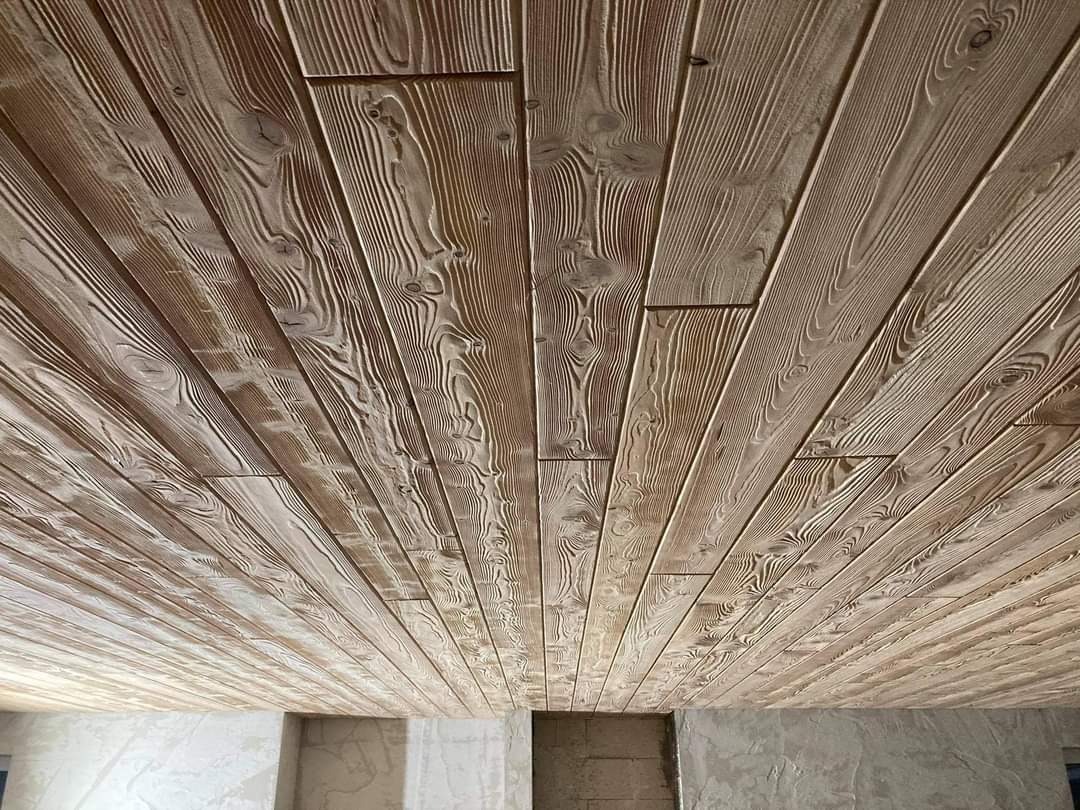 Drewniany sufit wyłożony jasnymi, lakierowanymi deskami z wyraźnym usłojeniem, widziany pod kątem, z fragmentem surowej ściany w tle.