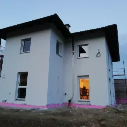 Dwukondygnacyjny dom z ocieploną elewacją i widocznymi rusztowaniami, uchwycony o zmierzchu, z widoczną różową izolacją fundamentów.