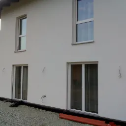 Świeżo otynkowana, biała elewacja domu jednorodzinnego z dwoma oknami i drzwiami balkonowymi, widoczne przygotowania do dalszych prac wykończeniowych w postaci rur kanalizacyjnych i kamiennego...