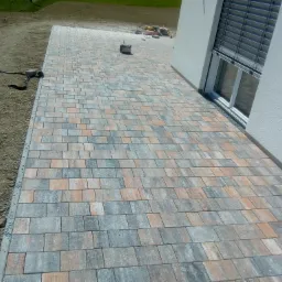 Ułożona kostka brukowa w odcieniach szarości i pomarańczy przylegająca do ściany budynku z oknem z żaluzjami zewnętrznymi; widoczne narzędzia i materiały budowlane w tle.