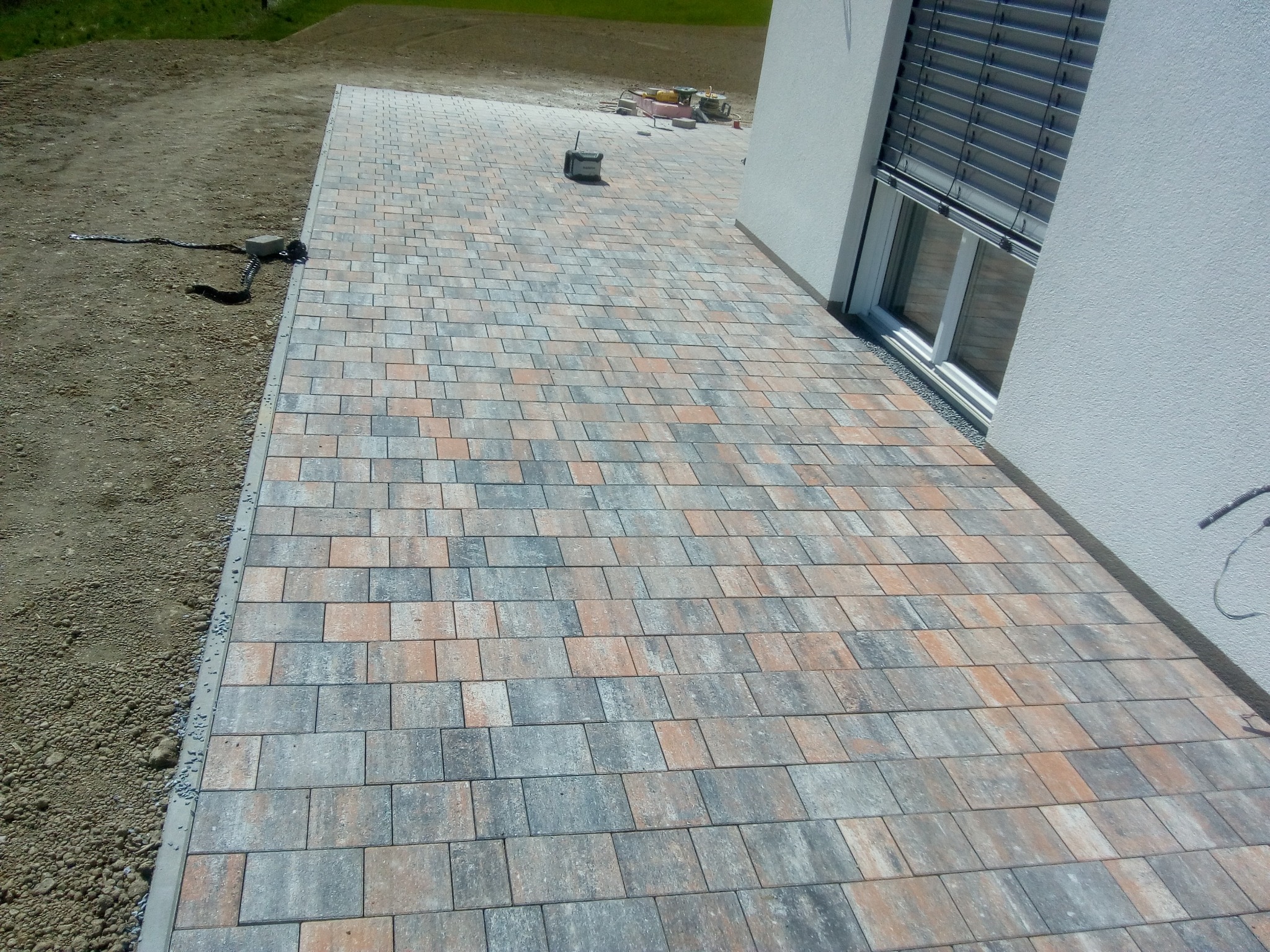 Ułożona kostka brukowa w odcieniach szarości i pomarańczy przylegająca do ściany budynku z oknem z żaluzjami zewnętrznymi; widoczne narzędzia i materiały budowlane w tle.