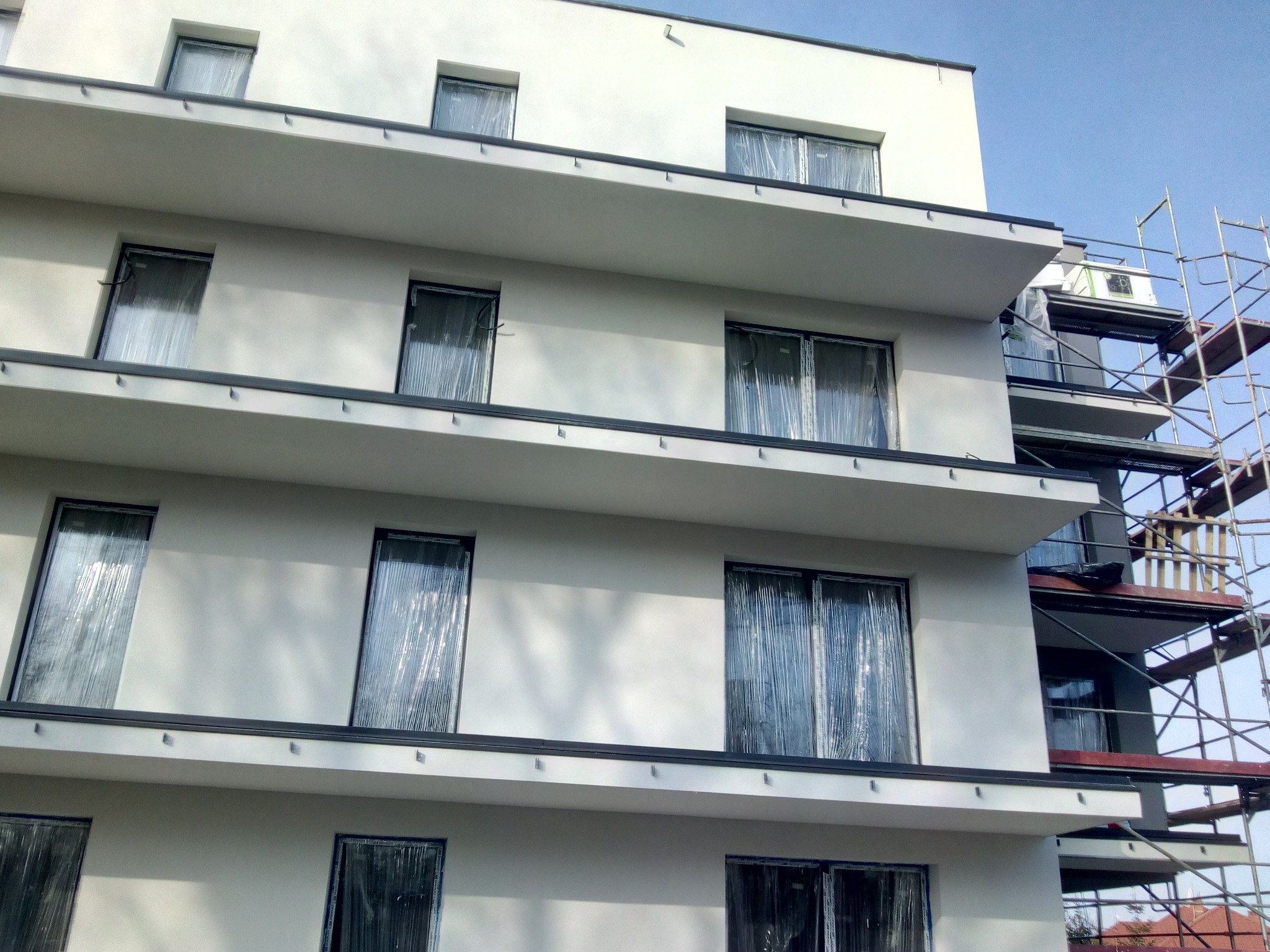 Nowoczesny budynek wielorodzinny z białymi ścianami i zabezpieczonymi folią oknami przed tynkowaniem, widoczne rusztowania.