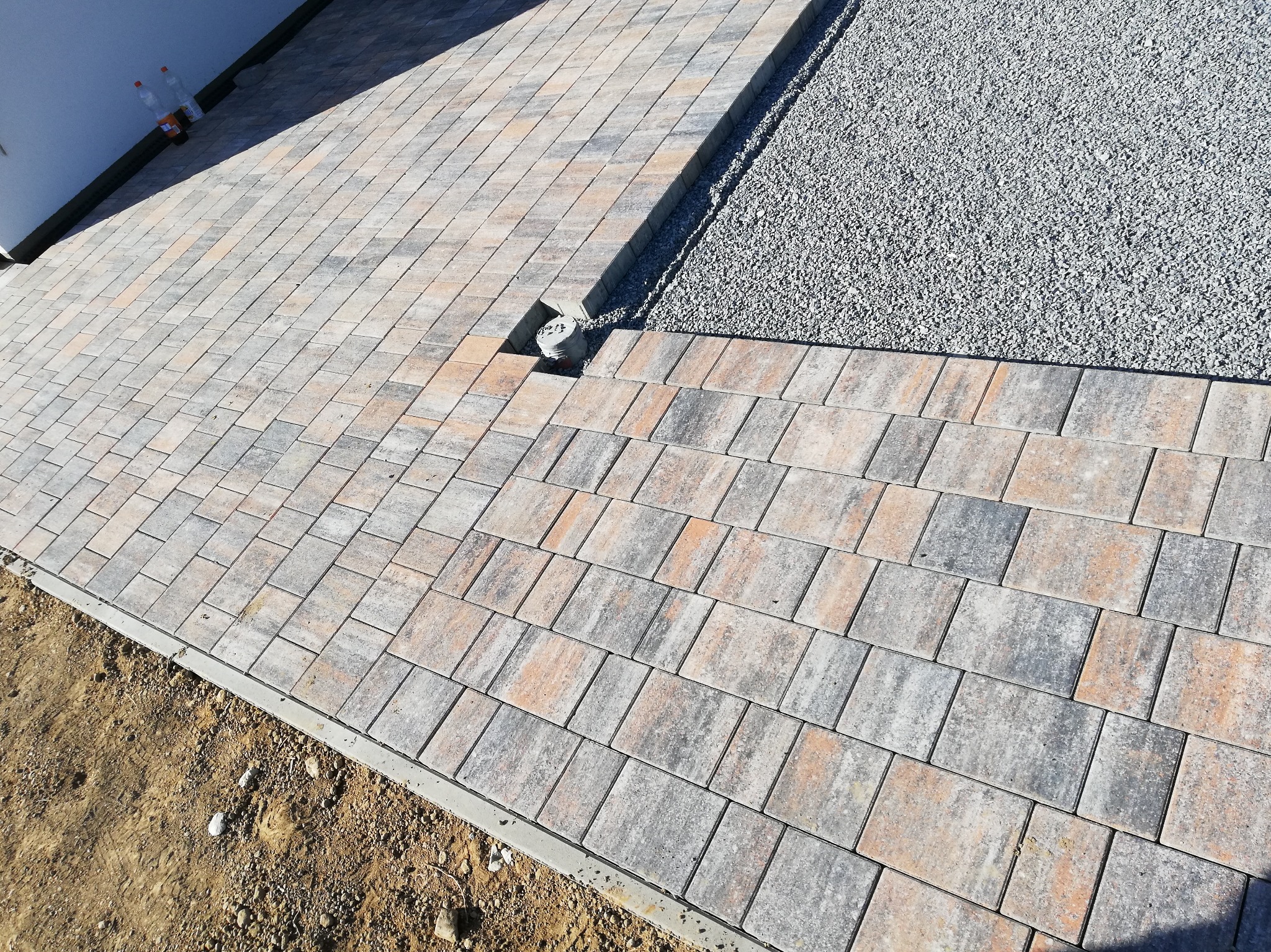 Ułożona kostka brukowa w odcieniach szarości i pomarańczy, z widocznym odpływem liniowym i fragmentem terenu z drobnym żwirem.