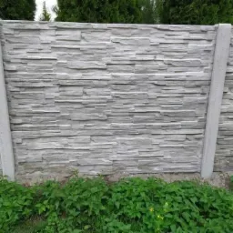 Szary, prefabrykowany płot z betonowych elementów imitujących kamień, z widocznymi słupkami i zielenią u podstawy.