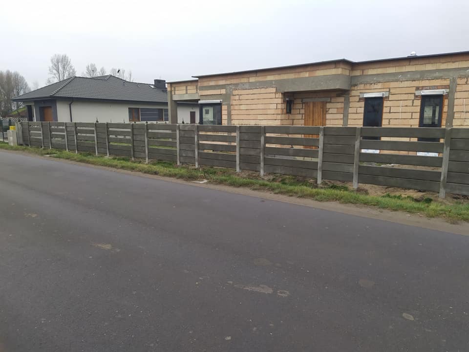 Szary, prefabrykowany płot z poziomych betonowych paneli między słupkami, oddzielający posesję z dwoma budynkami od asfaltowej drogi.