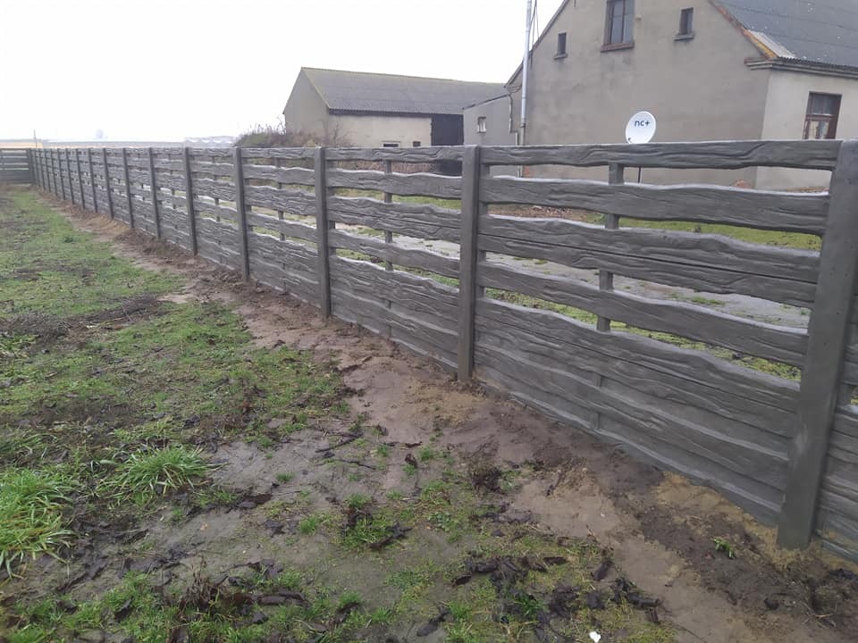 Długi, szary płot z prefabrykowanych elementów betonowych imitujących drewno, biegnący wzdłuż błotnistego terenu z rzadką trawą, z budynkami w tle i anteną satelitarną na jednym z nich.