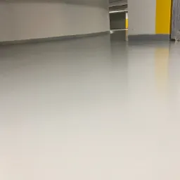 Jasnoszara, gładka posadzka w hali garażowej, widoczne żółte oznaczenia na ścianie i rury pod sufitem.