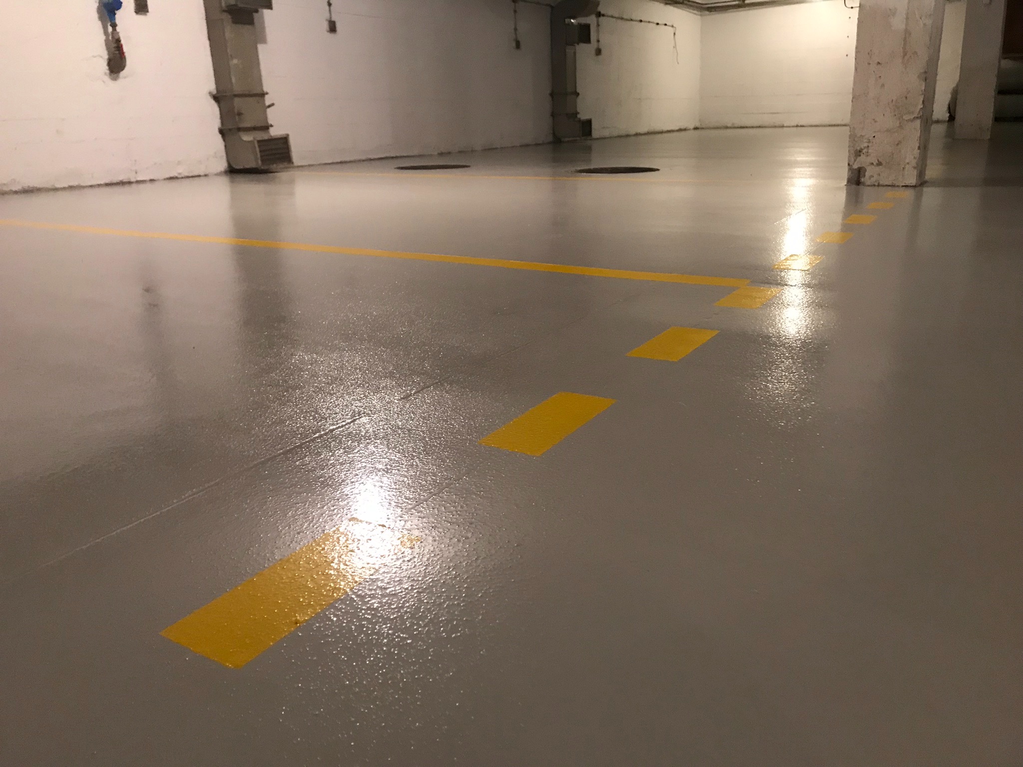 Szara, błyszcząca podłoga z żółtymi liniami i przerywanymi pasami w hali garażowej.