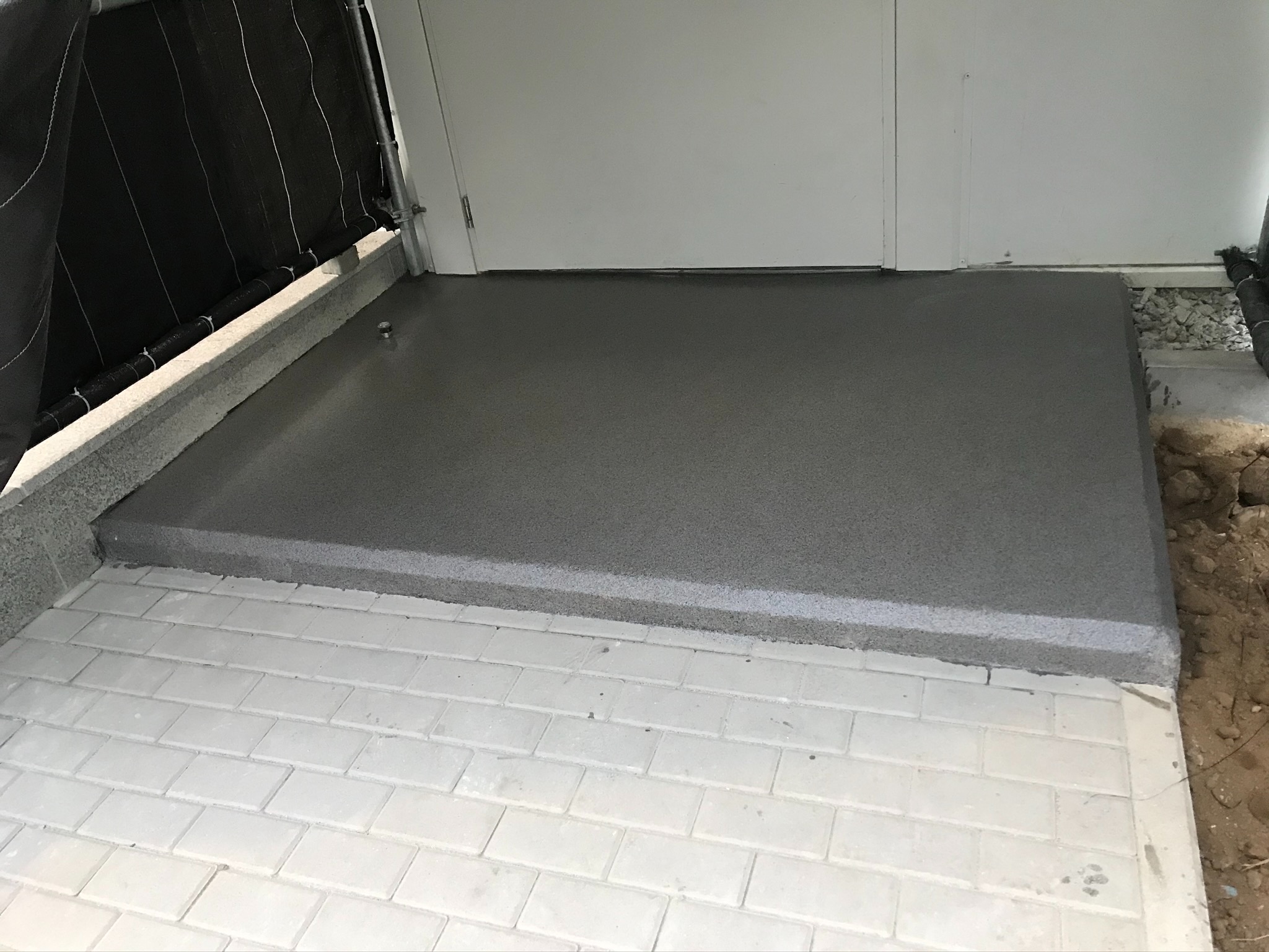 Świeżo wylana, szara, gładka powierzchnia w garażu, z widocznym fragmentem białej kostki brukowej przed wjazdem.