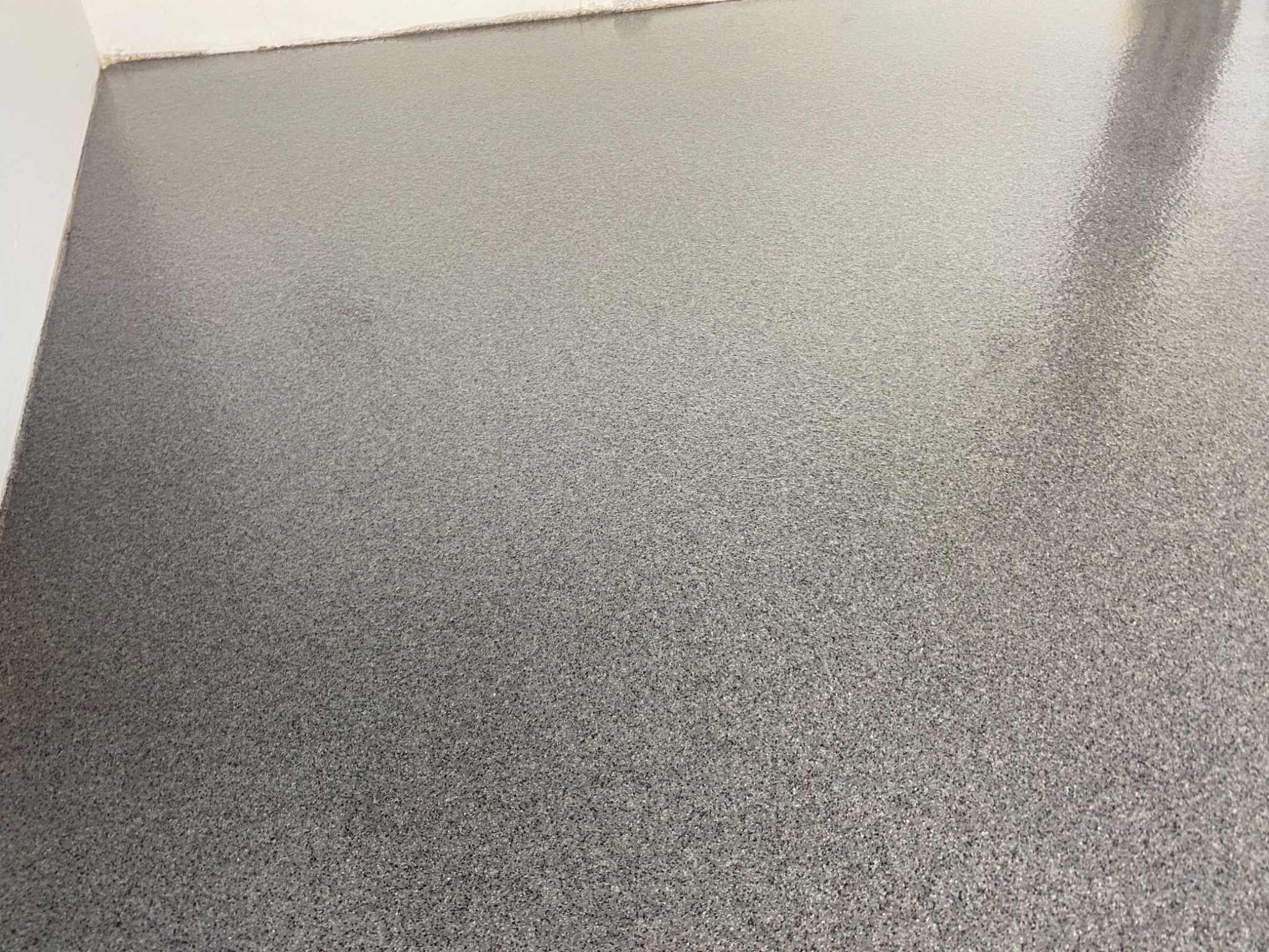 Szara, błyszcząca powierzchnia podłogi z drobnymi, ciemniejszymi wtrąceniami, widoczna przy białej ścianie.