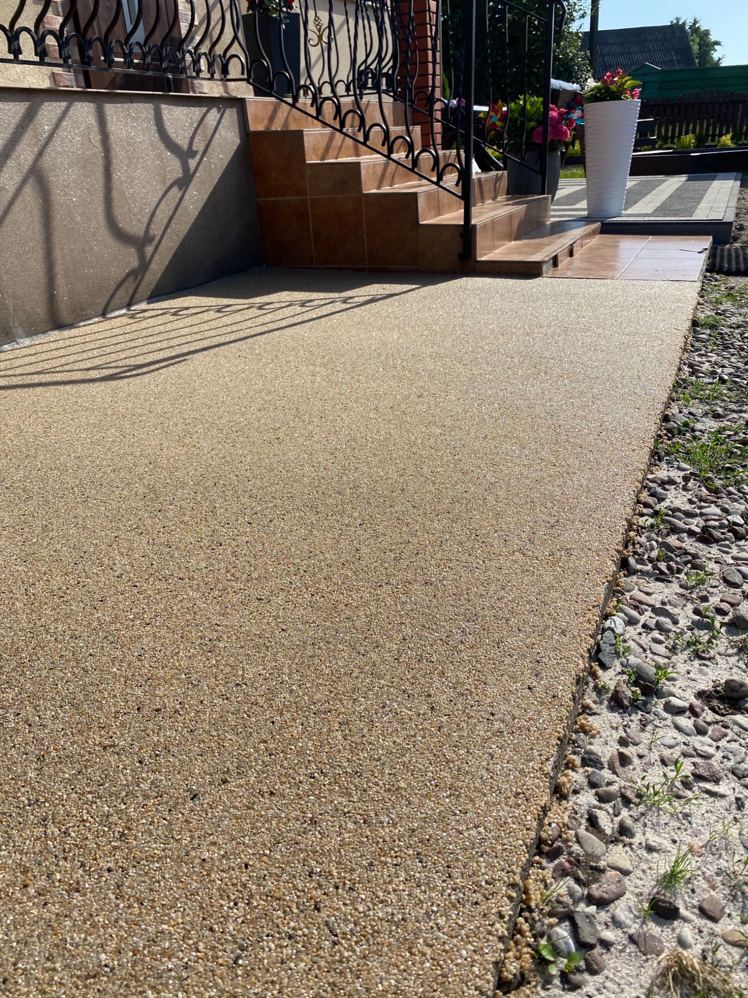 Beżowa powierzchnia z drobnych kamyczków prowadząca do schodów z brązowymi płytkami i czarną balustradą, obok rabata z kamieniami.