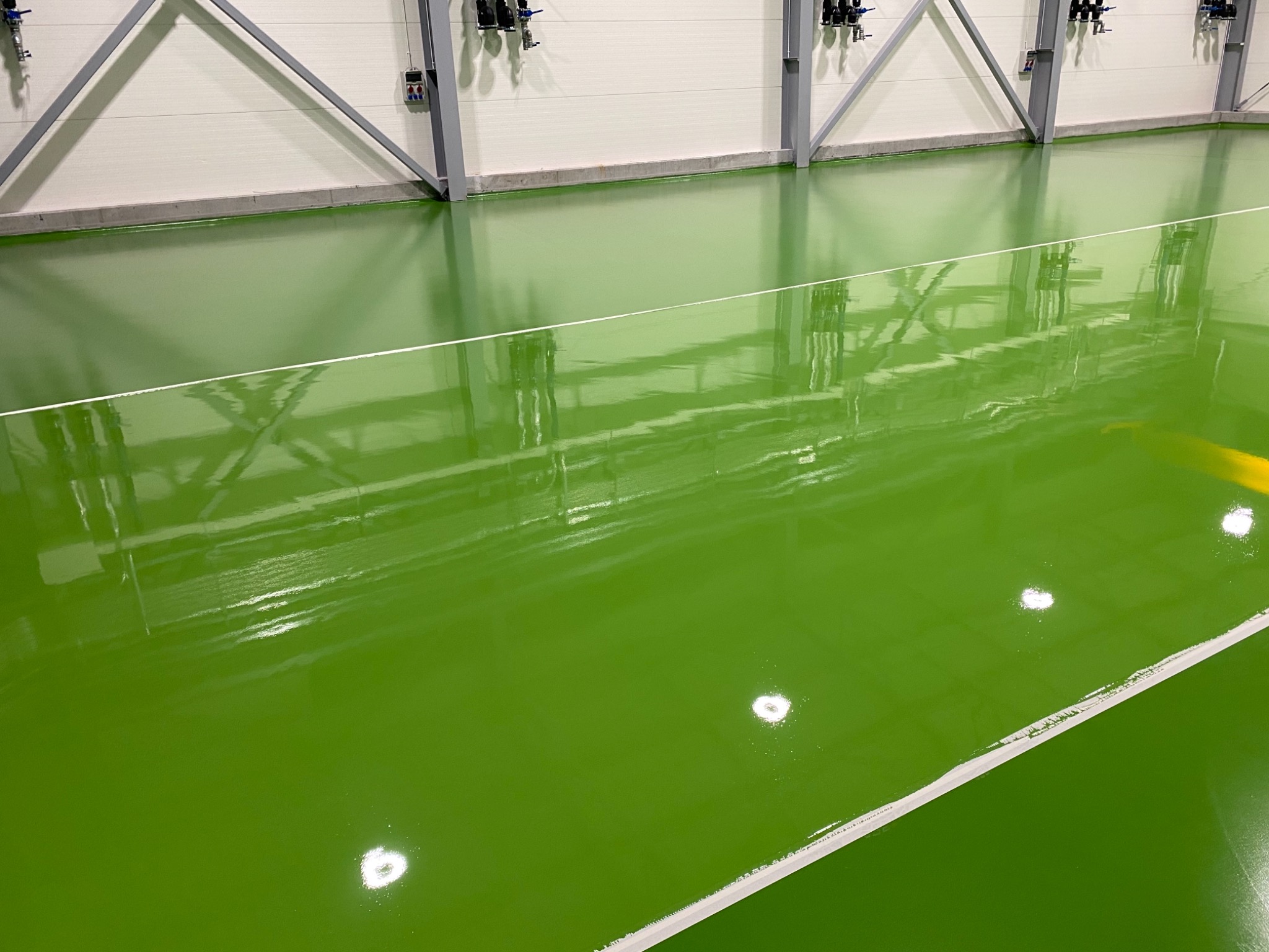 Jasnozielona, błyszcząca posadzka żywiczna w hali sportowej, z widocznymi białymi liniami boiska i odbiciami oświetlenia sufitowego.