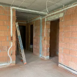 Surowy stan wnętrza z ceglanymi ścianami, widoczne przewody elektryczne poprowadzone po ścianach i suficie, oraz drabina oparta o ścianę. Podłoga betonowa, widoczne otwory drzwiowe.