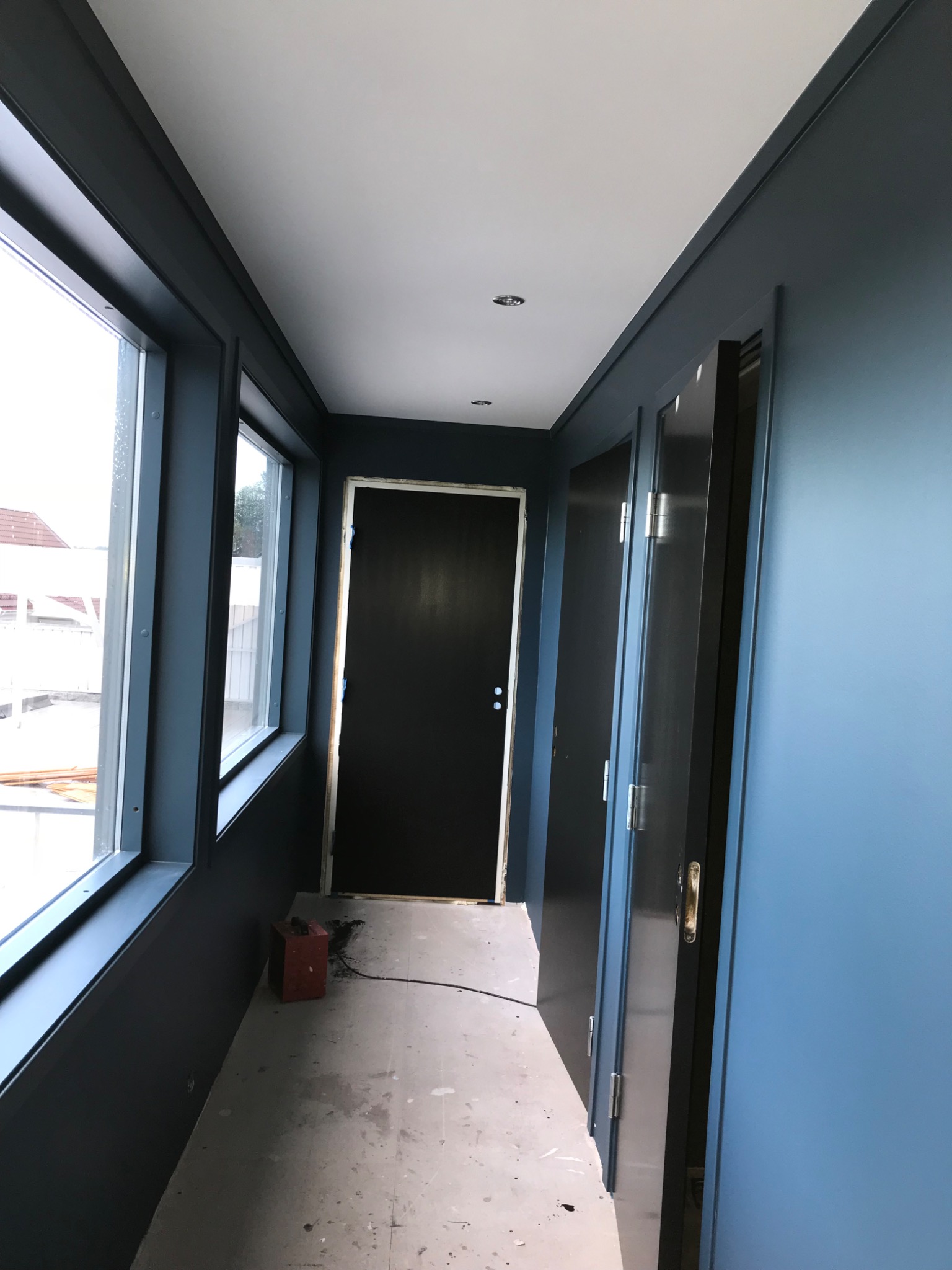 Korytarz w trakcie prac wykończeniowych, ściany pomalowane na ciemny odcień niebieskiego, widoczne okna z ciemnymi ramami, czarne drzwi i ślady farby na podłodze.