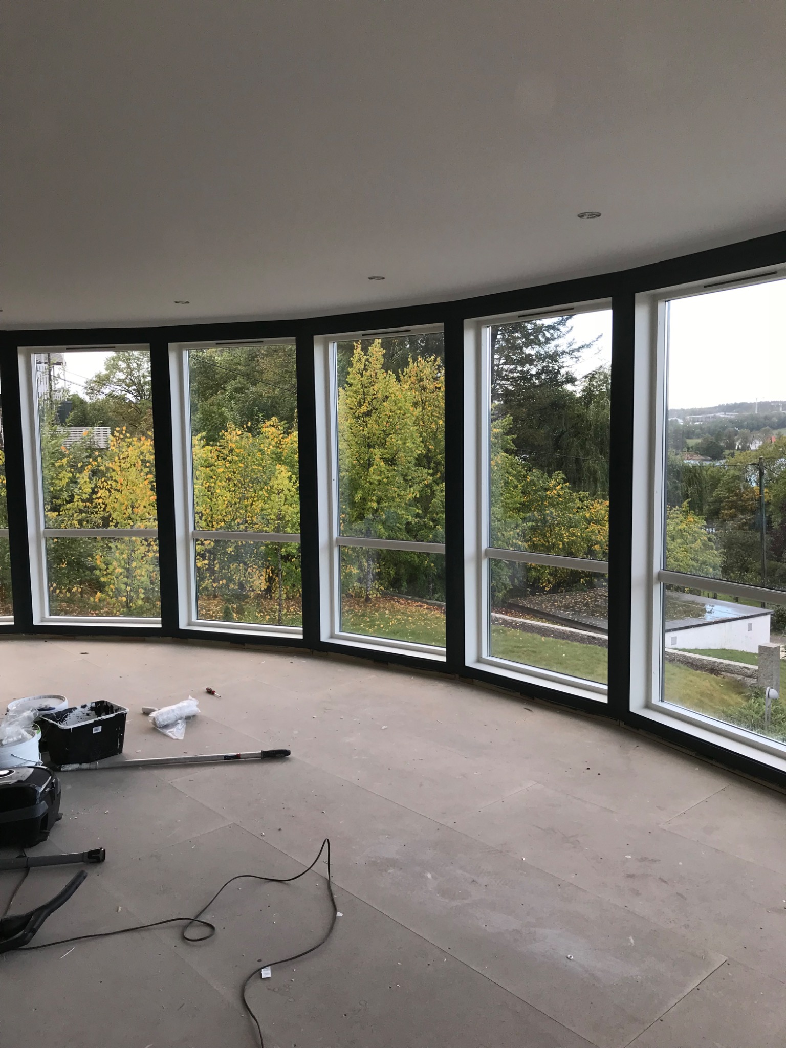 Przygotowanie do prac wykończeniowych w pomieszczeniu z zaokrągloną ścianą i dużymi oknami, widoczne narzędzia malarskie i szpachlarskie, podłoga zabezpieczona przed zabrudzeniem.