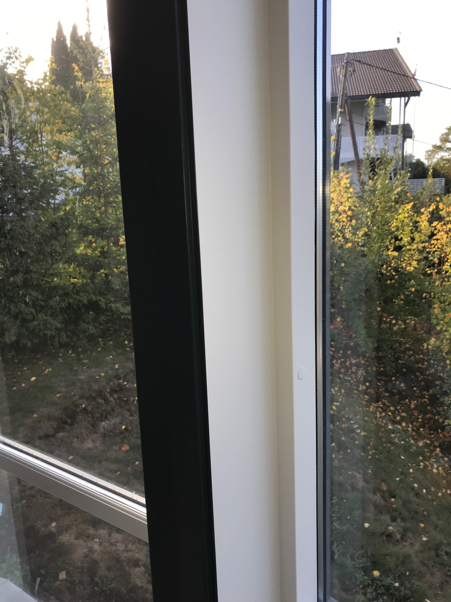 Nowe okno PCV z czarną ramą od wewnątrz, widok na ogród z drzewami i dom w tle.