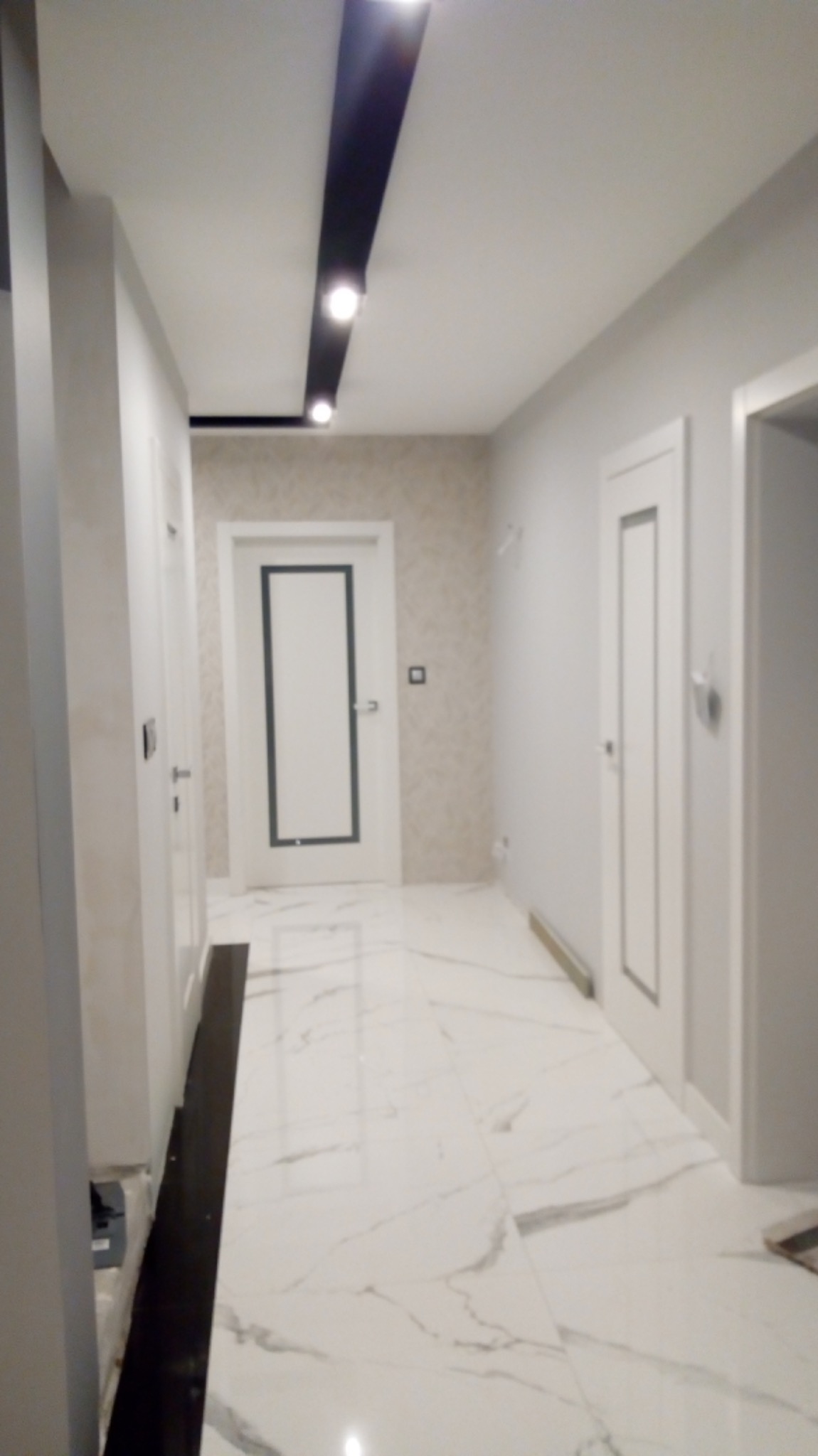 Korytarz w odcieniach szarości z marmurową posadzką, oświetleniem punktowym w czarnej listwie sufitowej i białymi drzwiami z szarymi obramowaniami.