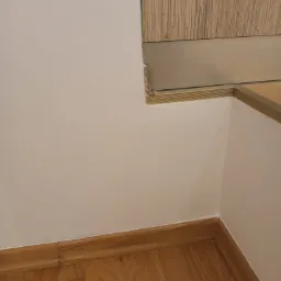 Ściana po malowaniu na biało, z drewnianą podłogą i listwami przypodłogowymi. Widoczny fragment schodów z drewnianym wykończeniem i tapetą na ścianie.