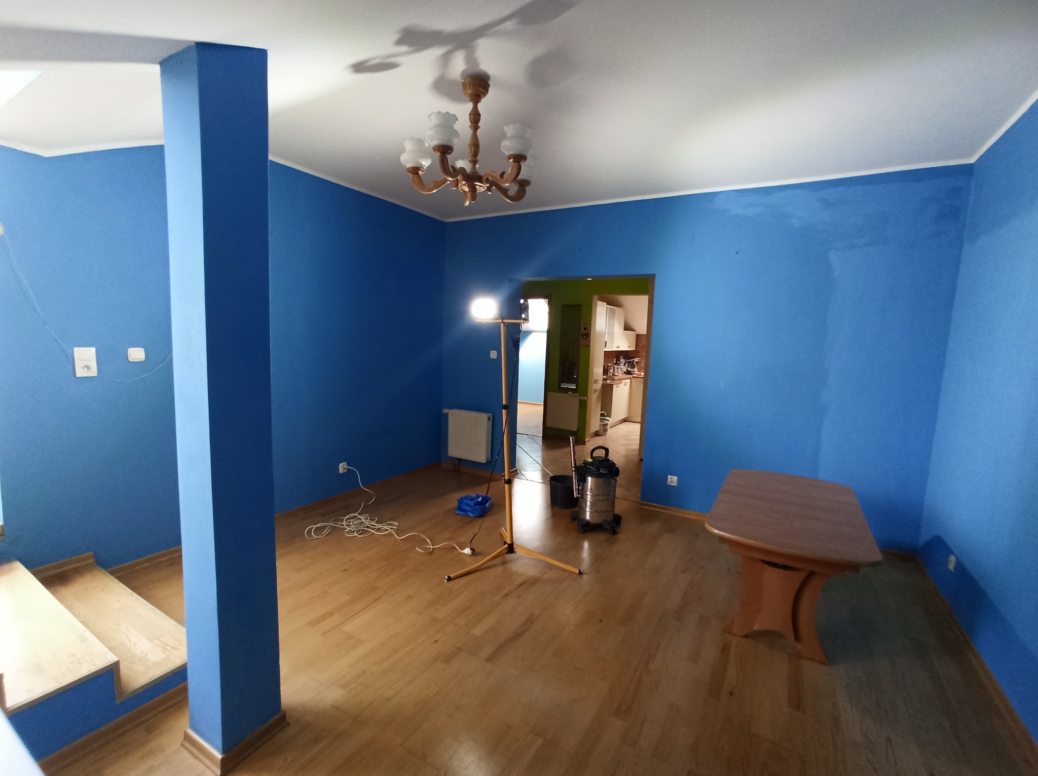 Pokój po malowaniu na niebiesko z lampą na statywie, odkurzaczem i stołem. Widoczne schody i fragment kuchni w tle. Świeżo pomalowane ściany, drewniana podłoga.
